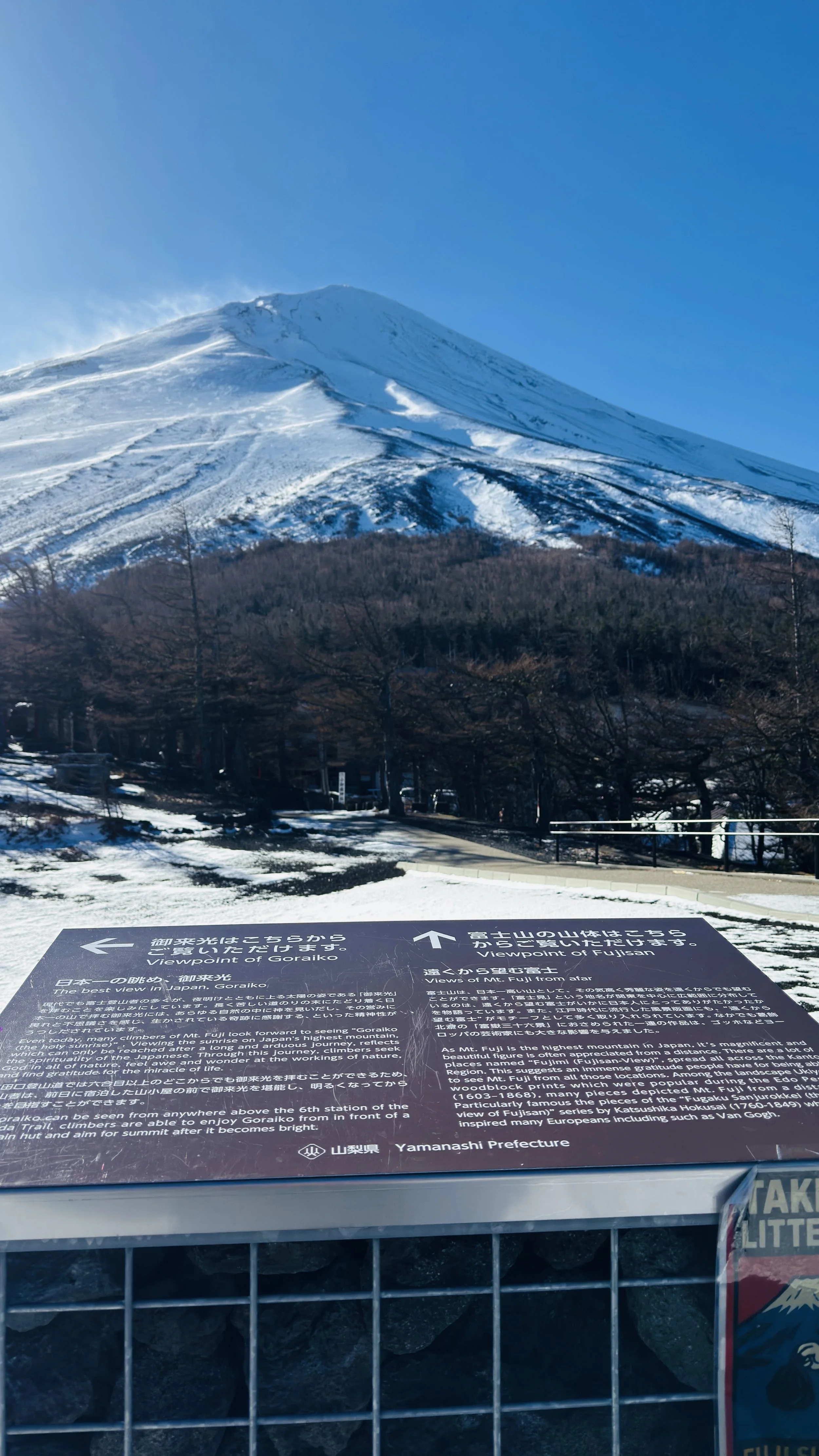 Snow-covered Mount Fuji against a clear blue sky, with a viewpoint sign in the foreground.