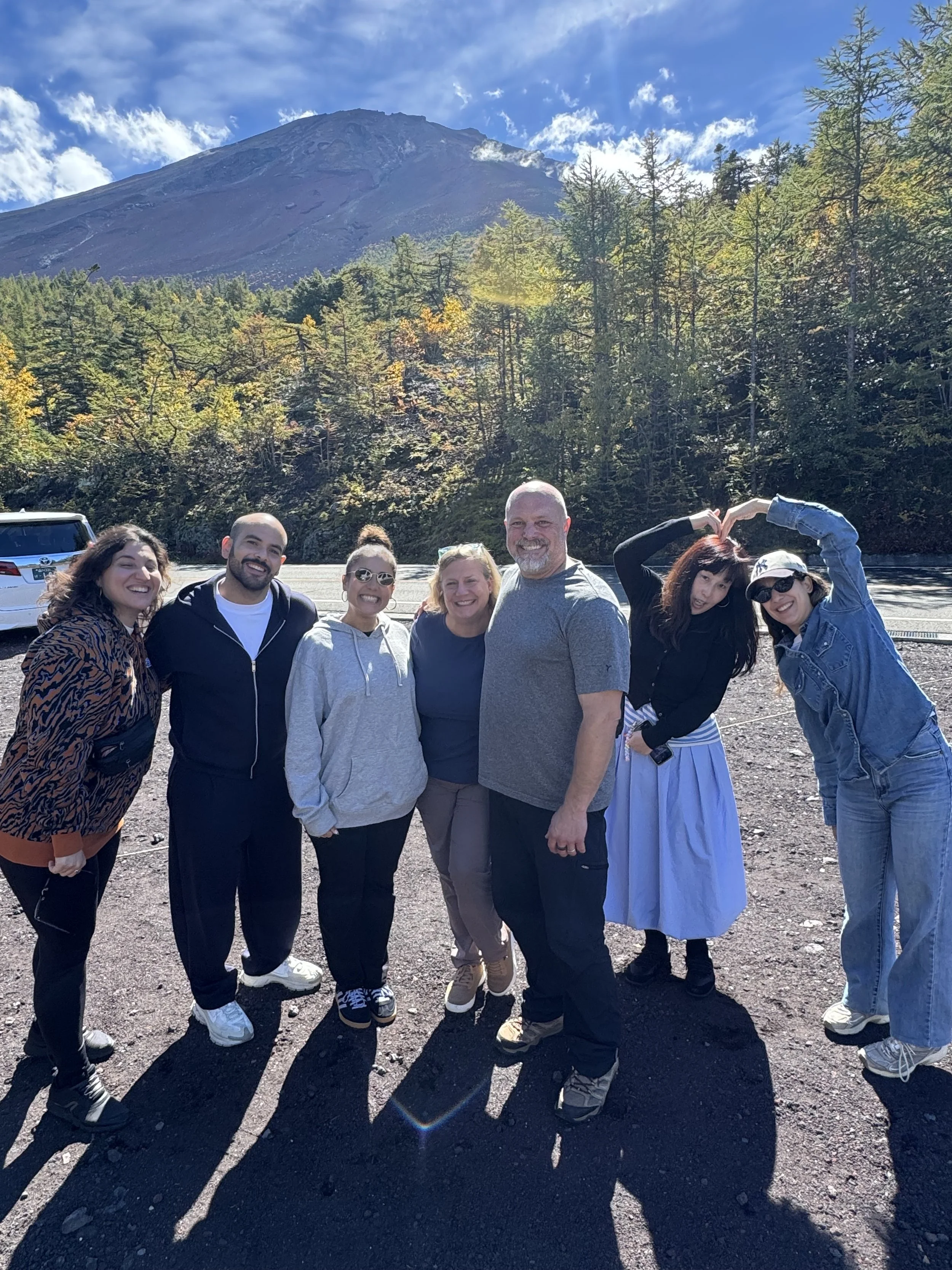 Group of seven people smiling and posing together outdoors with mountain and forest in the background.