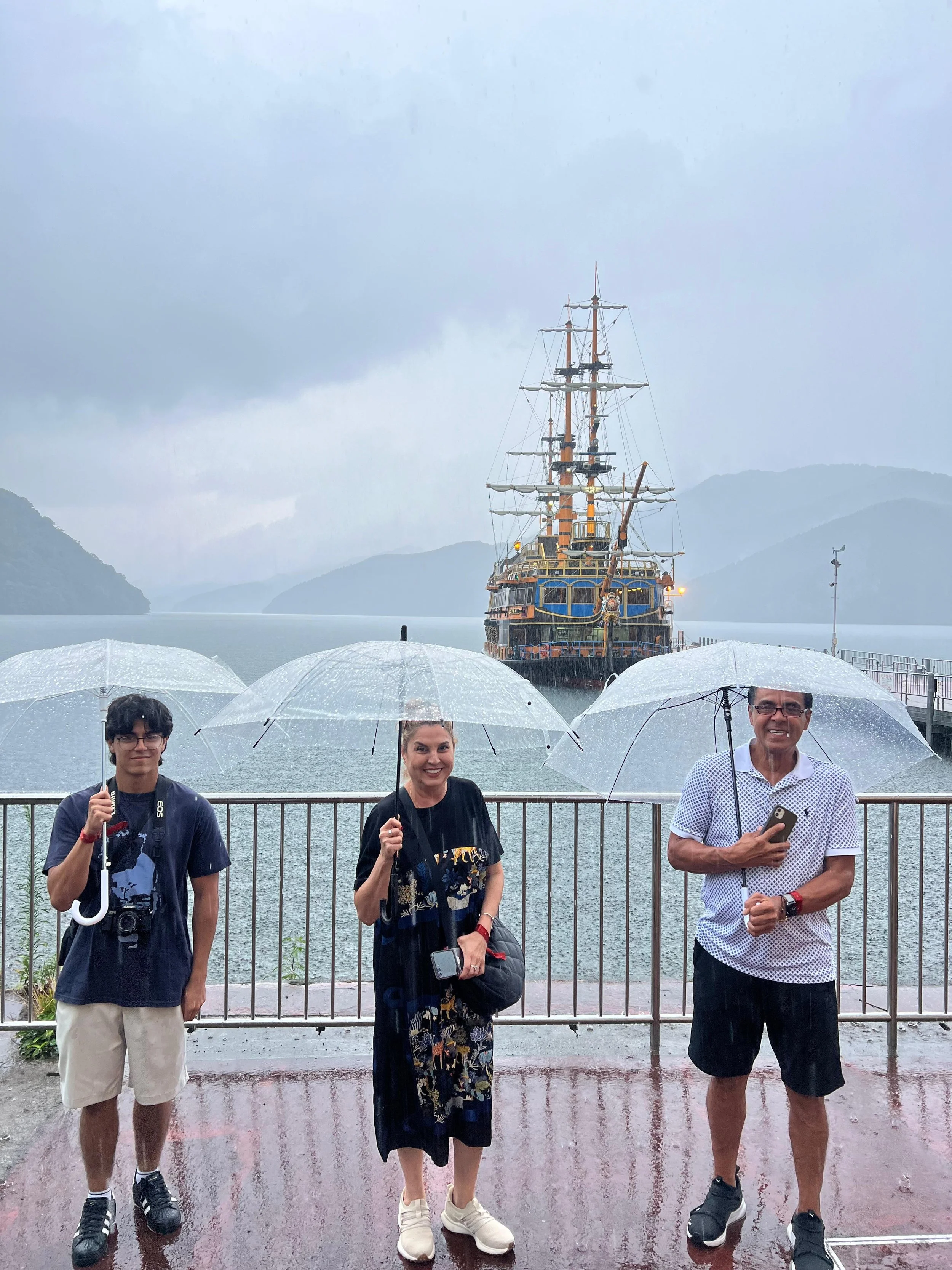 Three people standing on a dock holding umbrellas in the rain, with a large wooden pirate ship in the water and mountains in the background.