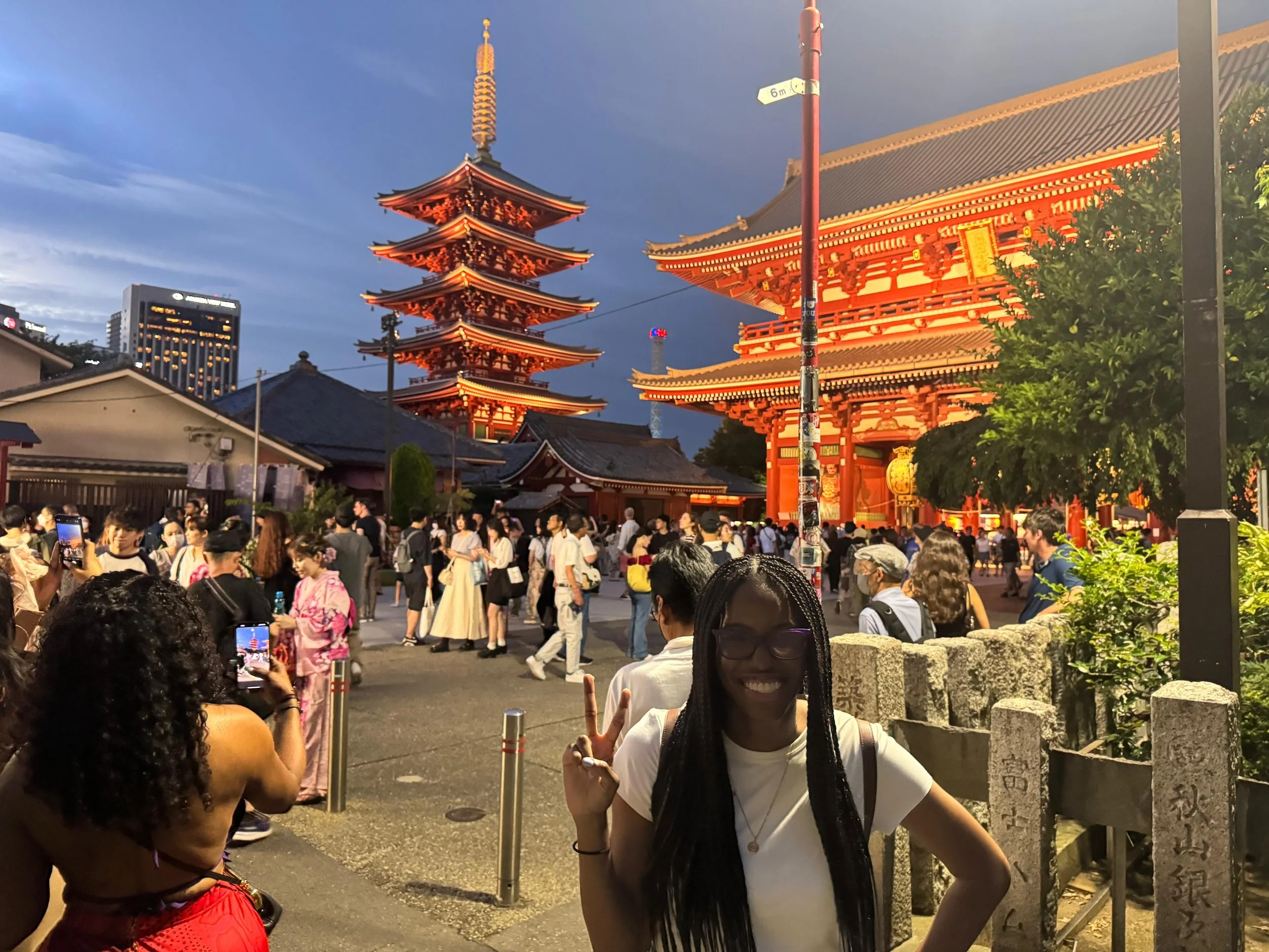 People posing for photos and walking outside a brightly lit traditional Japanese temple at dusk, with a five-story pagoda in the background and modern buildings nearby.