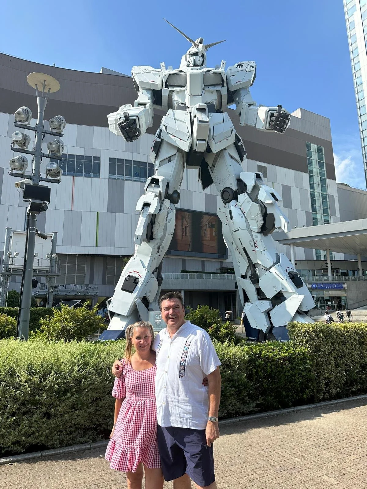 Two people, a woman in a pink checkered dress and a man in a white shirt and blue shorts, standing in front of a large Gundam statue outside a modern building. The man has his arm around the woman and they are smiling at the camera, with greenery and