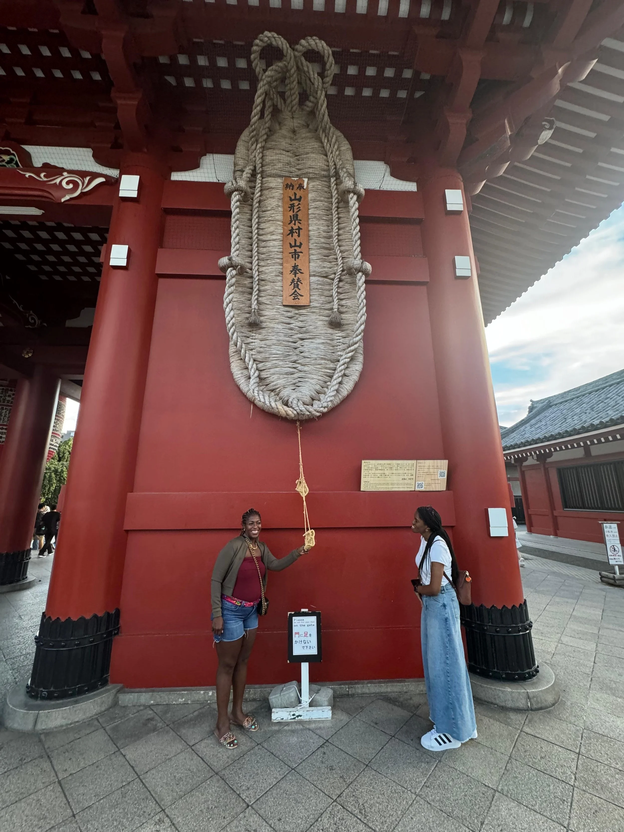 Two women standing under a large, hanging straw sandal at a temple. One woman is smiling and holding the sandal's rope, while the other woman is looking at her. The sandal has a wooden plaque with Japanese writing and is attached to a large red struc