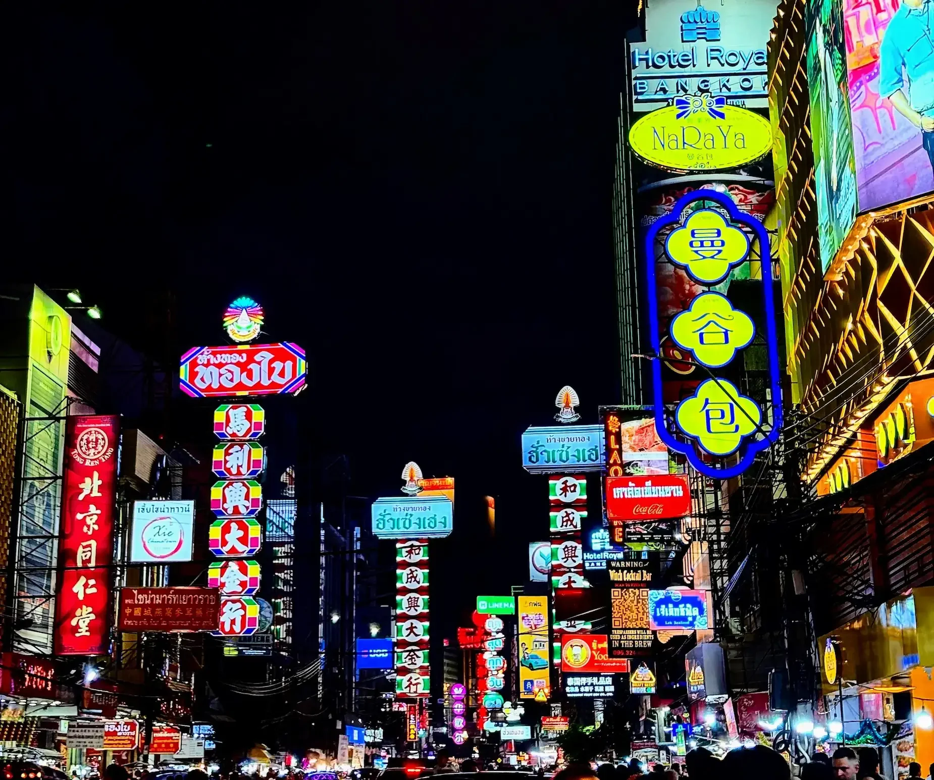 Night scene of a busy street in an Asian city, illuminated by numerous colorful neon signs and billboards in various languages, with pedestrians and vehicles below.