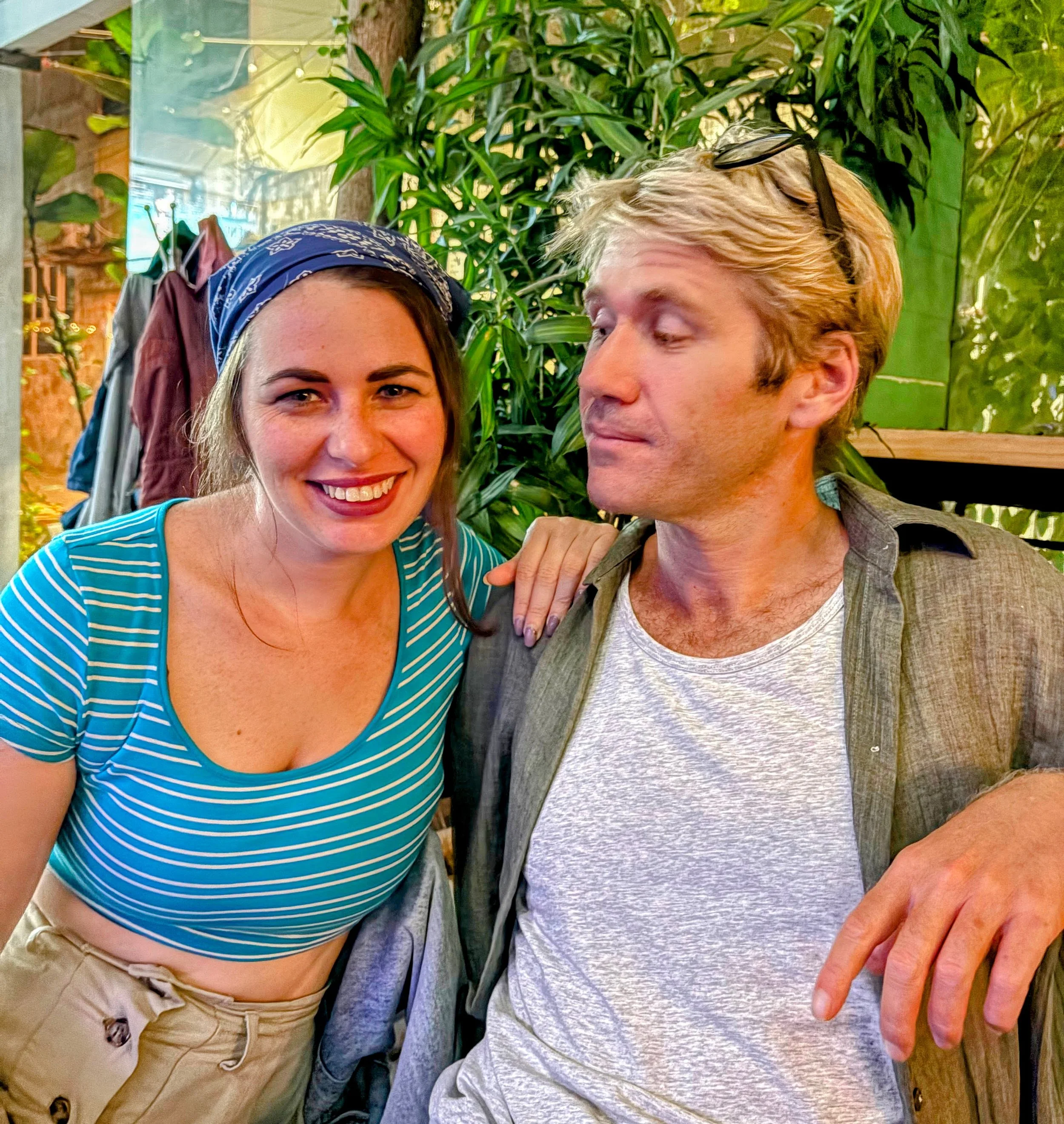 A smiling woman with long brown hair wearing a blue and white striped shirt and khaki pants, standing next to a man with blonde hair, glasses on his head, wearing a grey t-shirt and a green jacket, in a lush indoor setting with green plants.