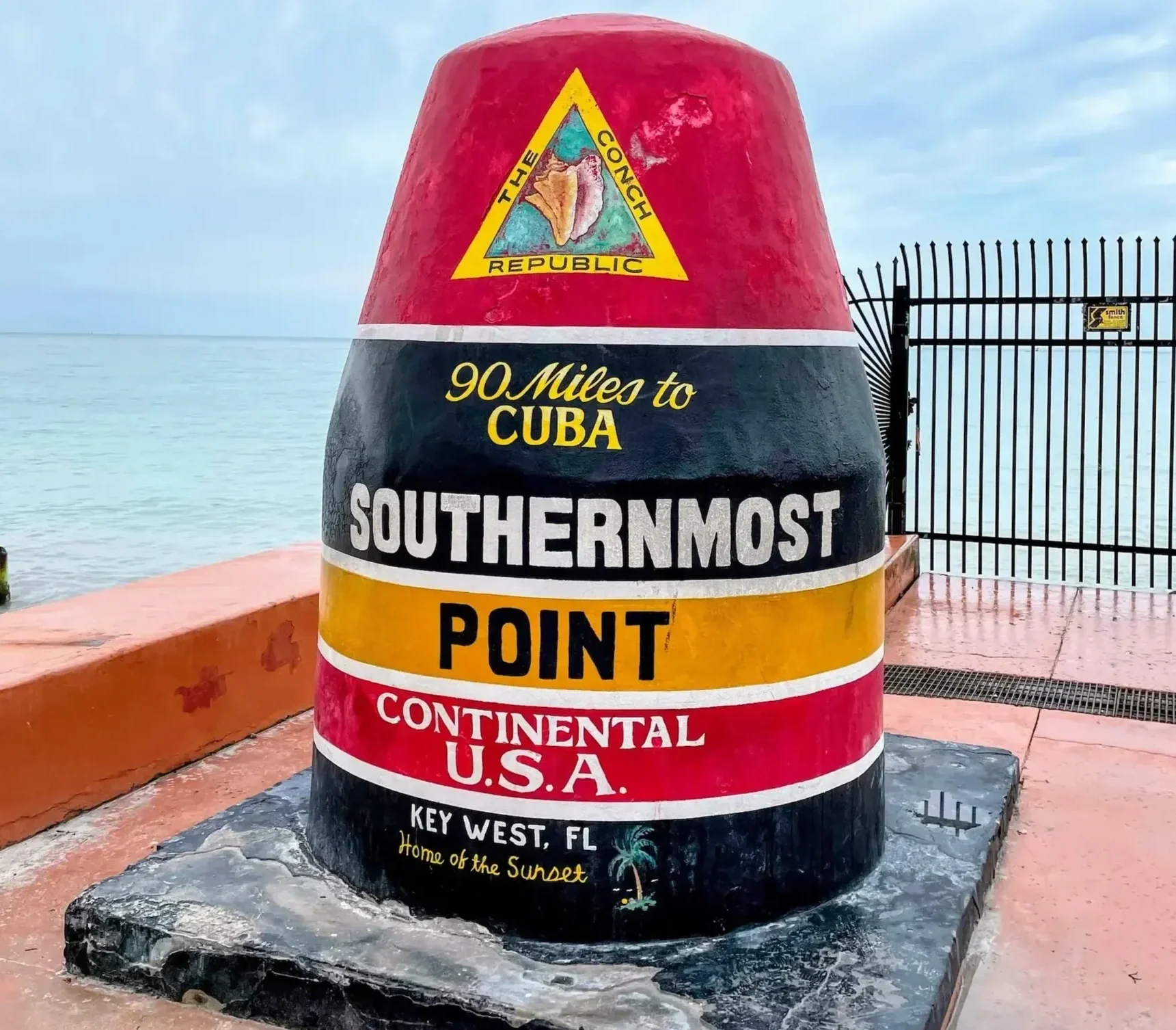 Colorful buoy marker at Key West, Florida, indicating the southernmost point of the continental United States, with the ocean in the background and a fence to the right.
