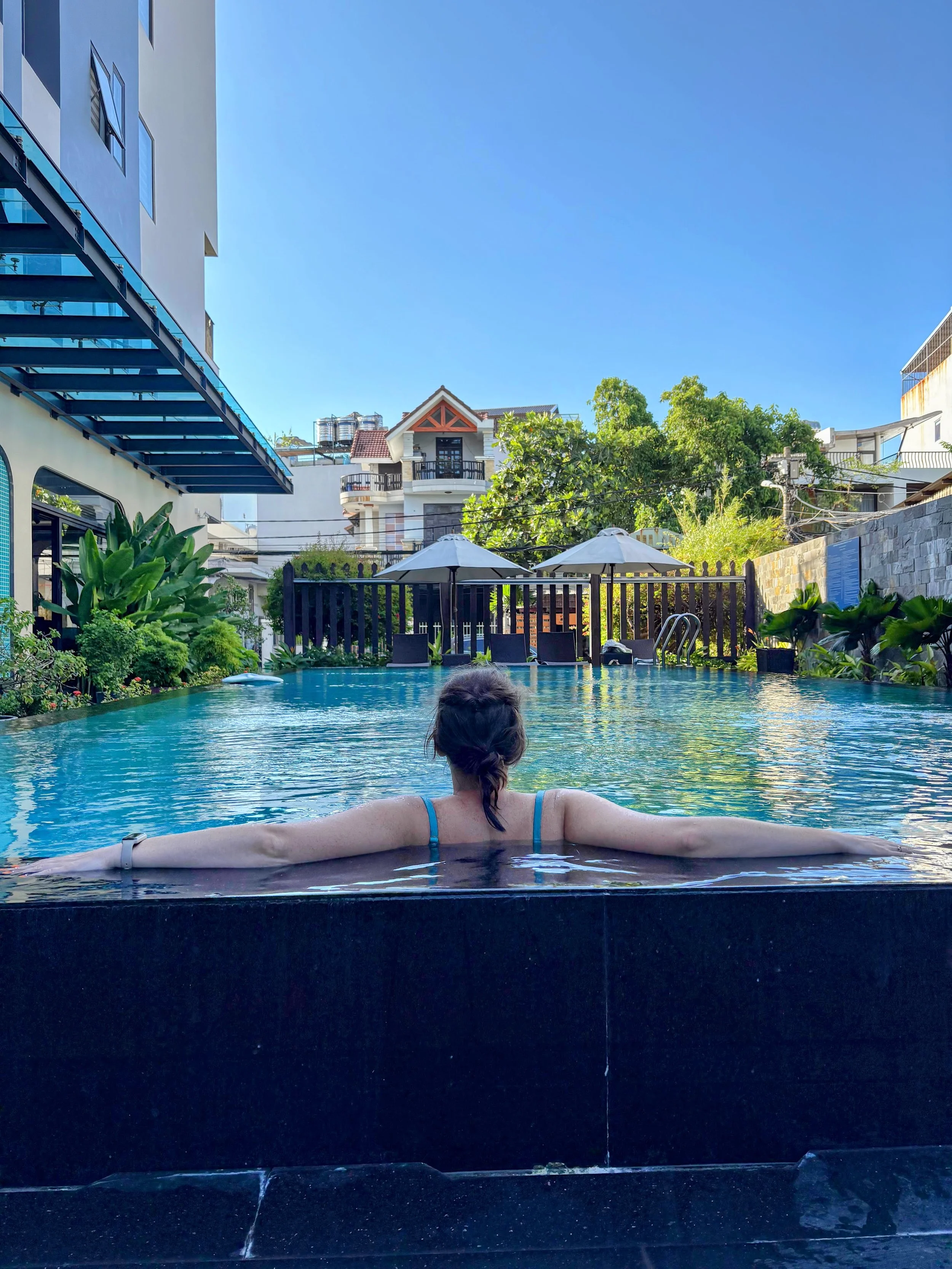 A woman swimming in a pool with her back to the camera, surrounded by greenery and residential buildings, under a clear blue sky.