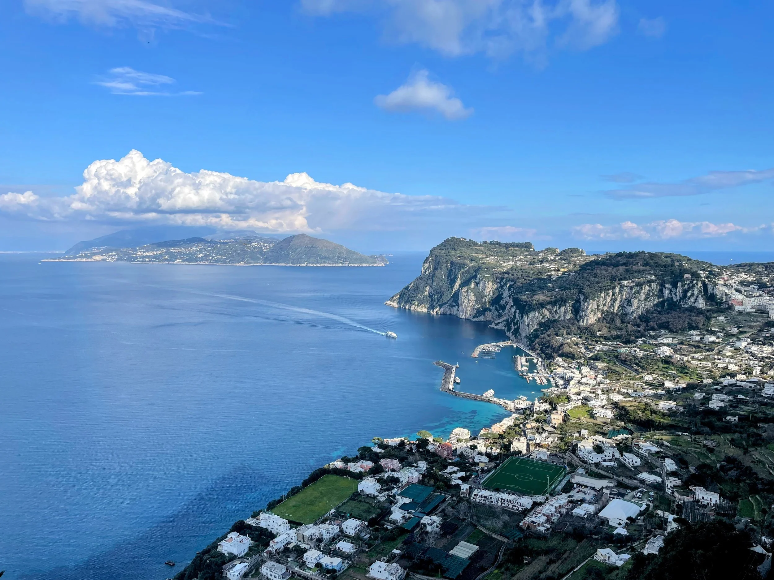 Aerial view of a coastal town with surrounding cliffs and mountains, ocean with a boat and marina, and a sports field in the foreground.