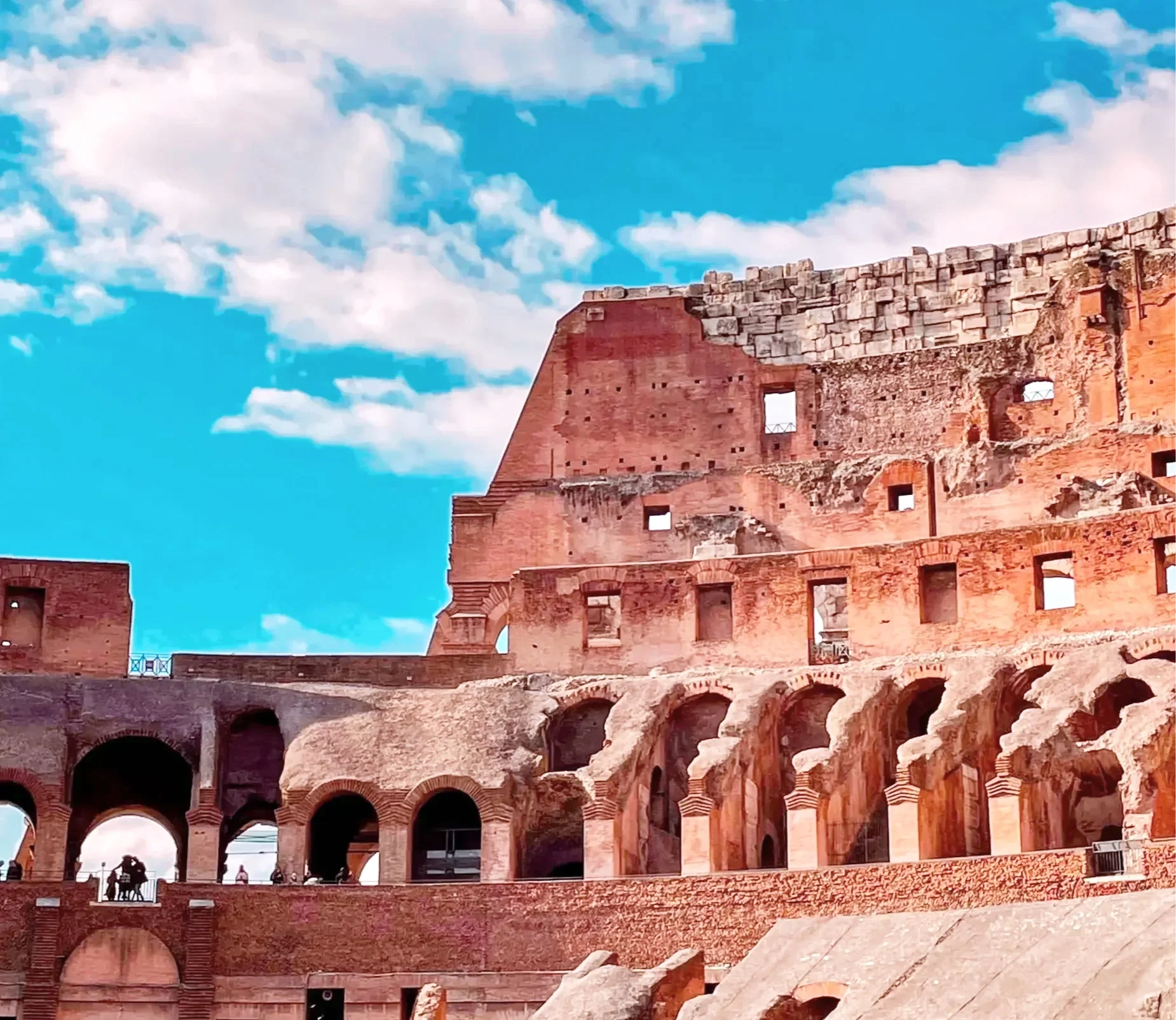The image shows a partial view of the Colosseum in Rome, Italy. The ancient amphitheater's arched brick and stone structure is visible against a bright blue sky with scattered white clouds. There are people near the base of the structure, enjoying the view.