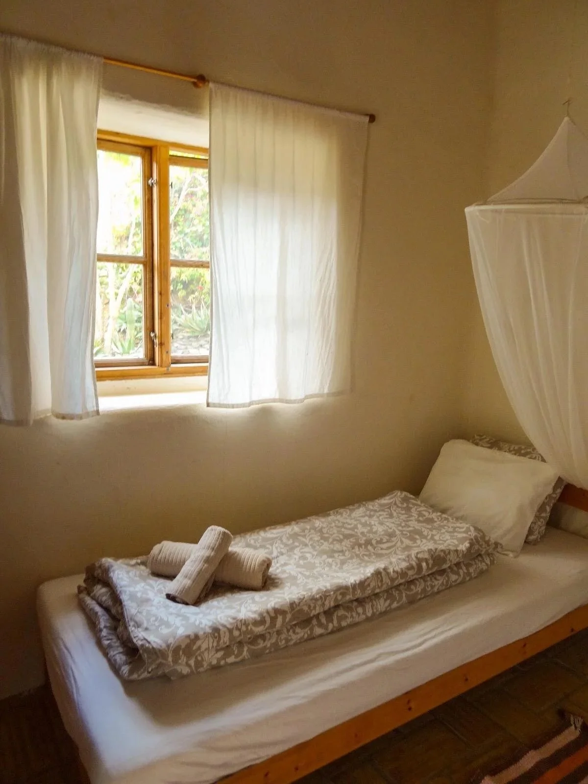 A small bedroom with a single bed, white bedding, a pillow, and a mosquito net. There is a window with wooden framing and white curtains, letting in natural light.