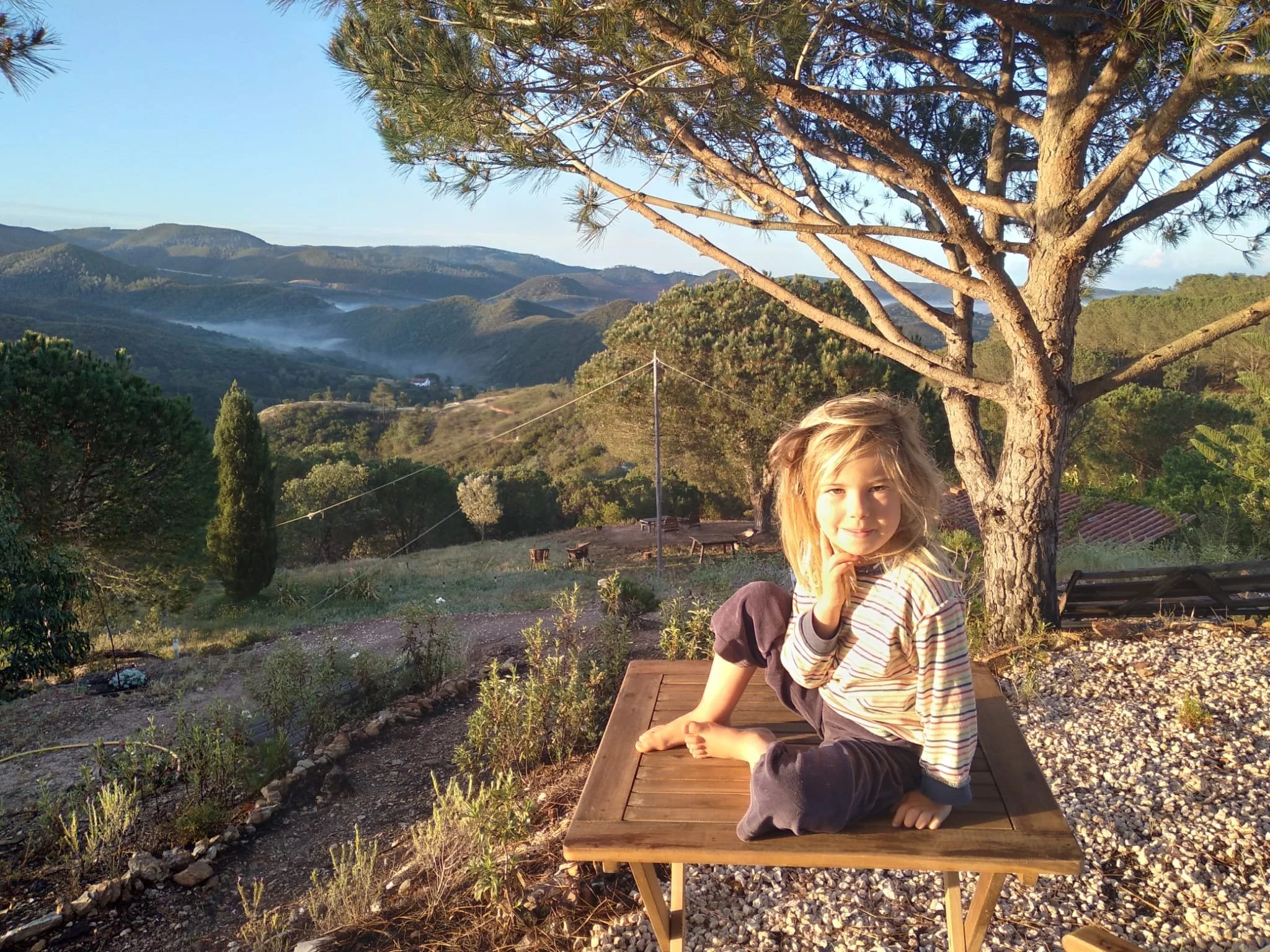 A young girl sitting on a wooden table outdoors in front of a tree, with mountains and a valley in the background during sunset.
