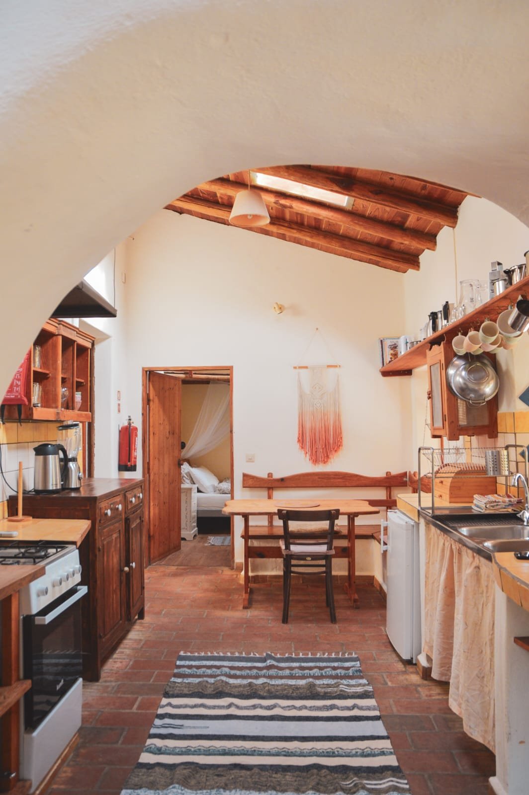 Cozy rustic kitchen with wooden cabinets, a small dining table, and views into a bedroom with a bed and canopy.