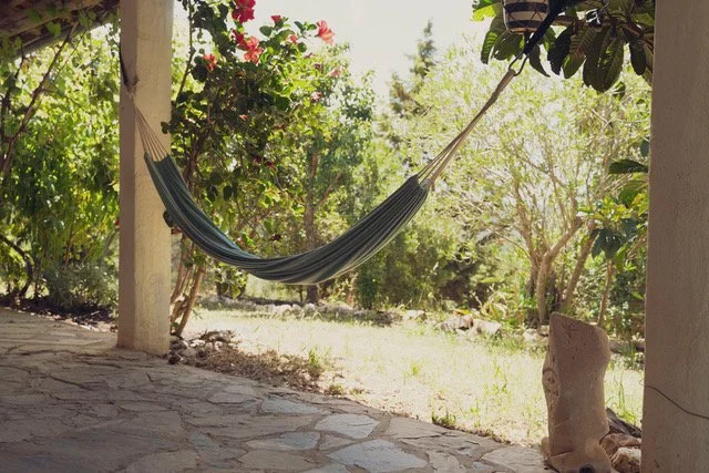A hammock hanging between two porch posts on a stone patio with trees and sunlight in the background.