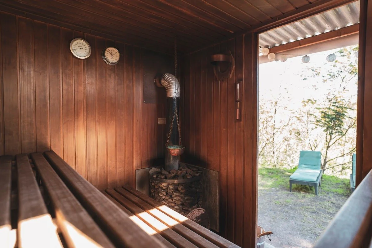 Wood-paneled sauna room with a wooden bench, two climate control gauges on the wall, a pipe connected to a stove, and an open door leading outside to a grassy area with outdoor chairs.