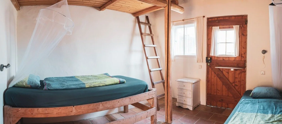 Room with a bunk bed, another bed, a small wooden door, a window, and a ladder. The room has wooden accents and tile flooring.