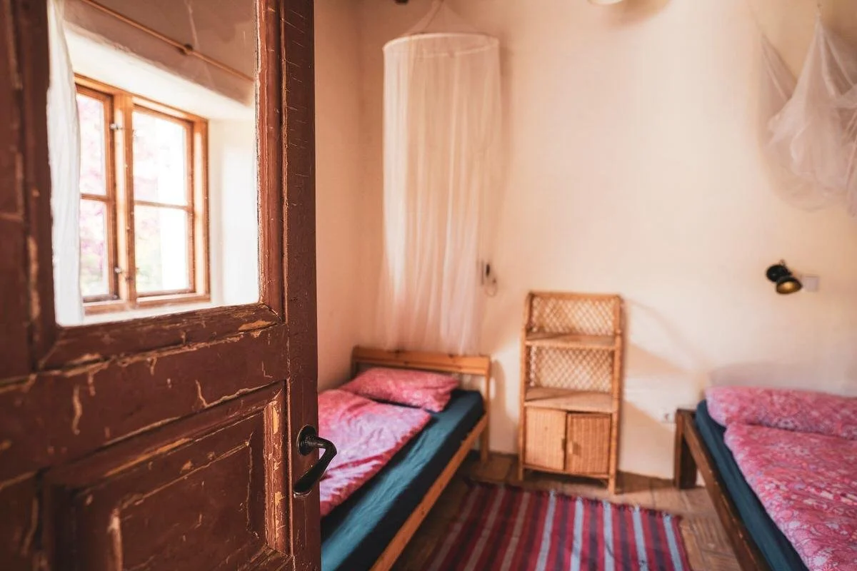 A cozy bedroom with two beds covered in pink floral blankets, a wooden chair, a small cabinet, and a window with wooden framing letting in natural light.