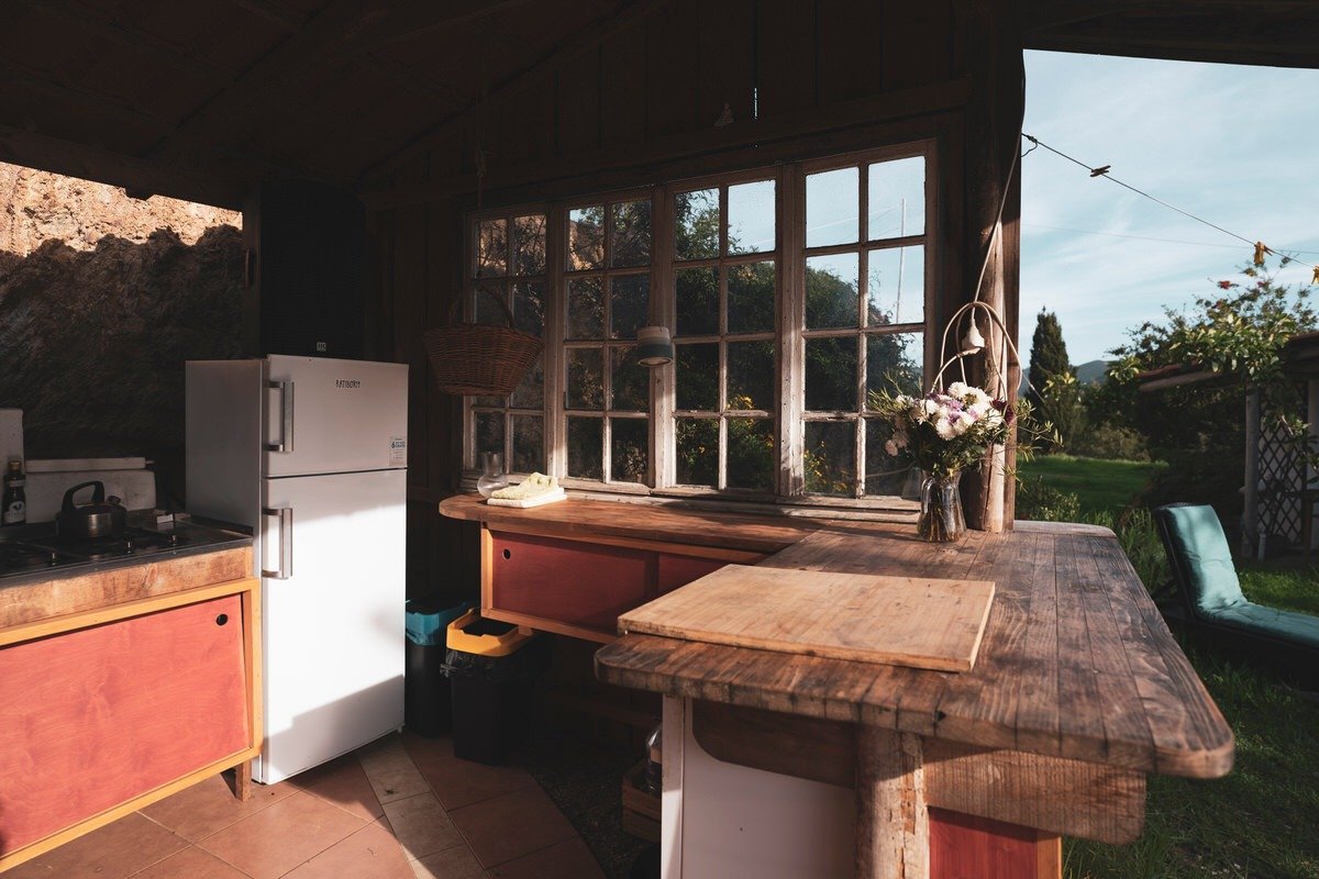 A rustic kitchen with a wooden countertop, a white refrigerator, and a window overlooking a garden with greenery and trees.