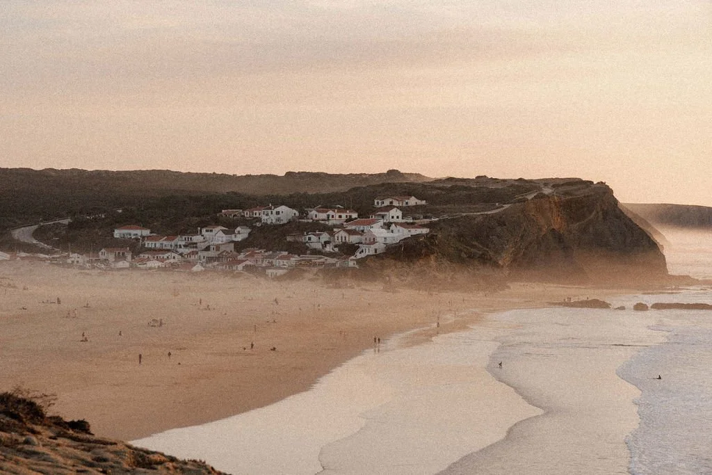 A beach scene with people walking on the sand and surfing in the water. A cliff with a cluster of white houses is visible in the background, with the ocean waves crashing against the shore at sunset.