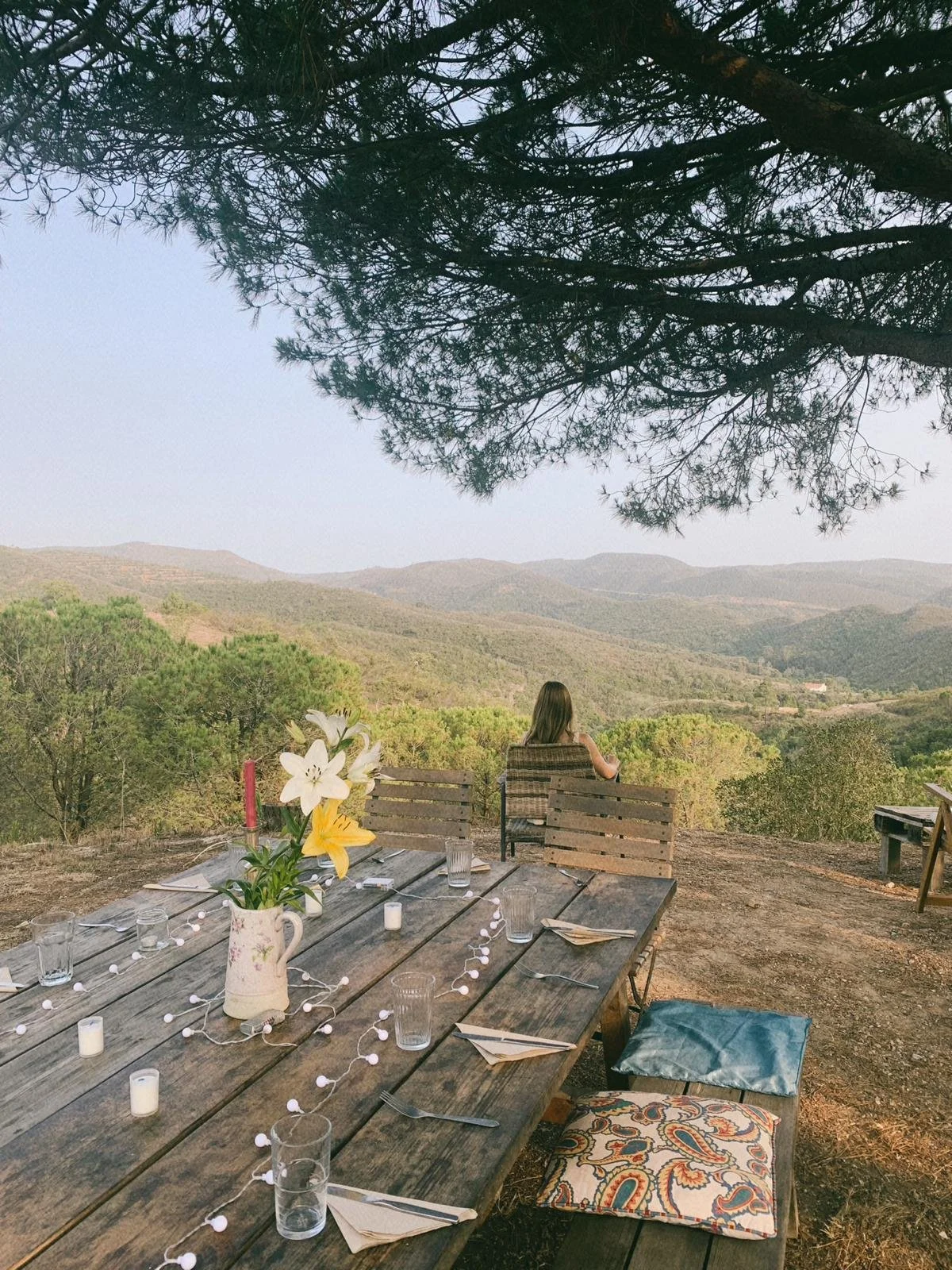 Outdoor dining table set for a gathering with flower centerpiece and string lights, overlooking a scenic mountain landscape with a woman sitting on a bench.