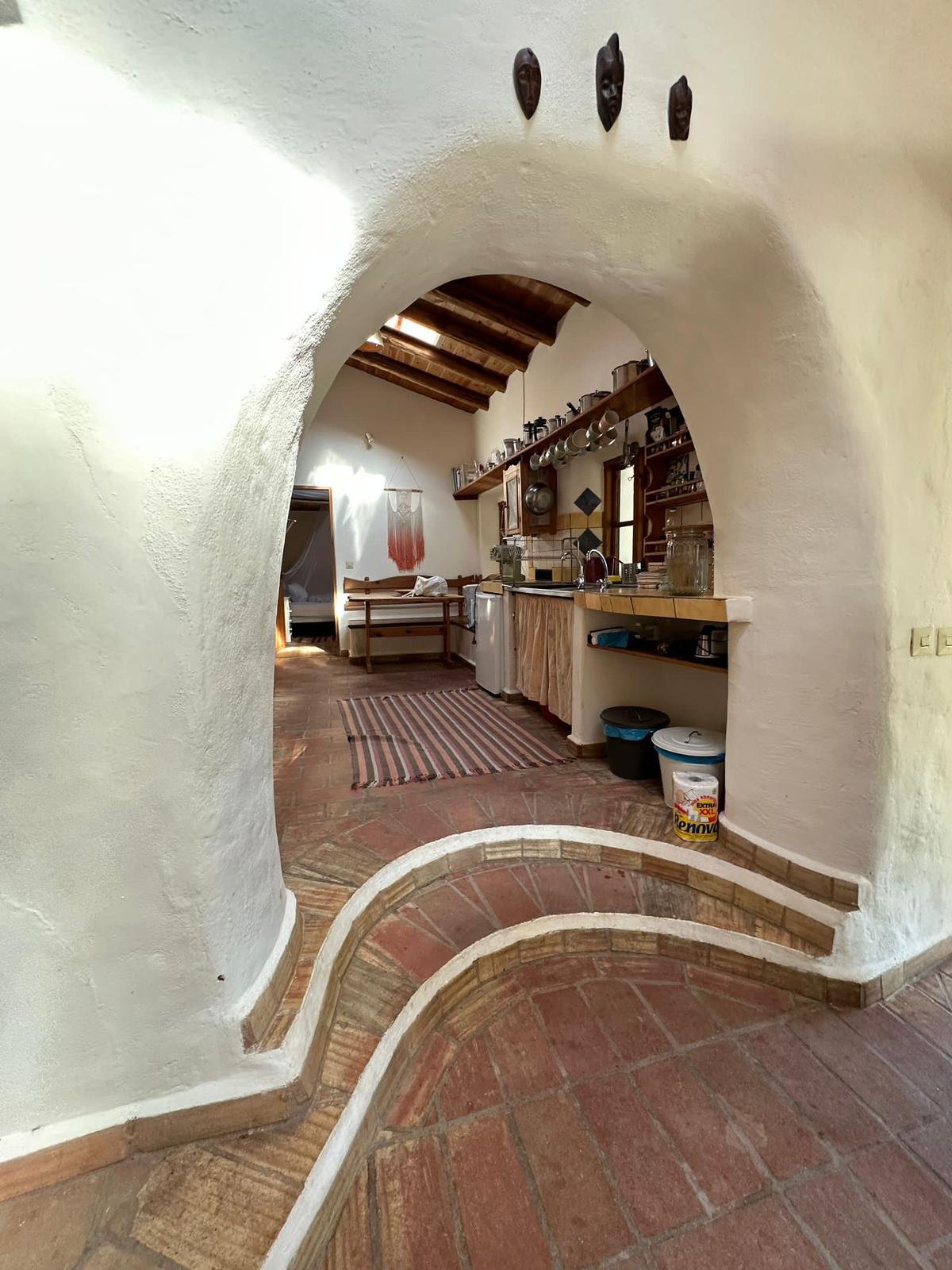 View of a rustic kitchen and dining area with terracotta tile flooring, white textured walls, and an arched entryway with steps. The kitchen has wooden cabinets, open shelving with dishes and glassware, a small island, and a window with sunlight. Dec
