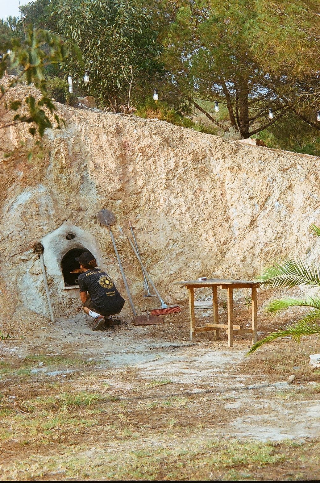 Person wearing black t-shirt and cap crouching by small oven or kiln in outdoor setting, with brooms and a table nearby, surrounded by trees and string lights.