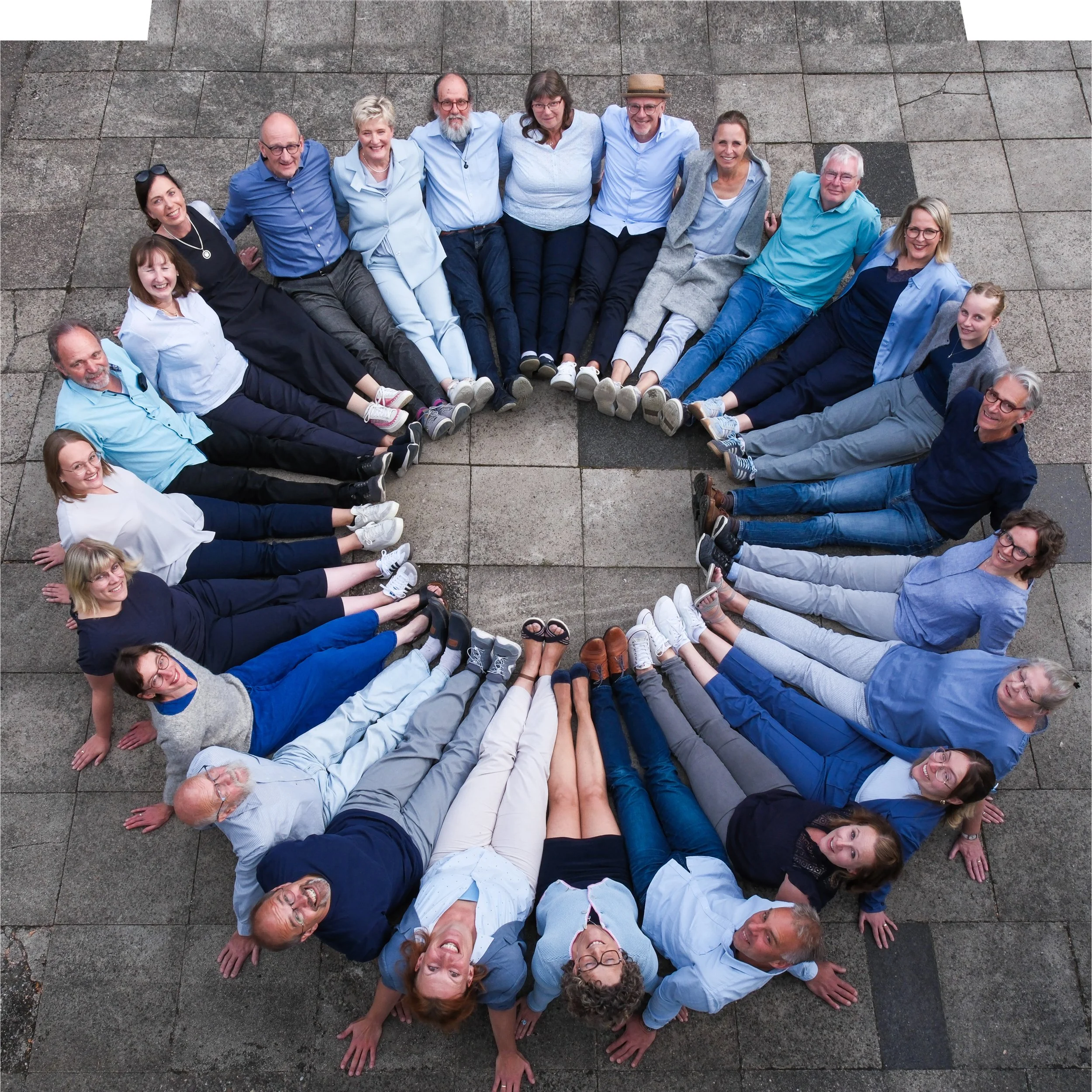Menschen sitzen im Kreis auf einem Steinboden, schauen nach oben, lachen und haben die Beine zusammengelegt.
