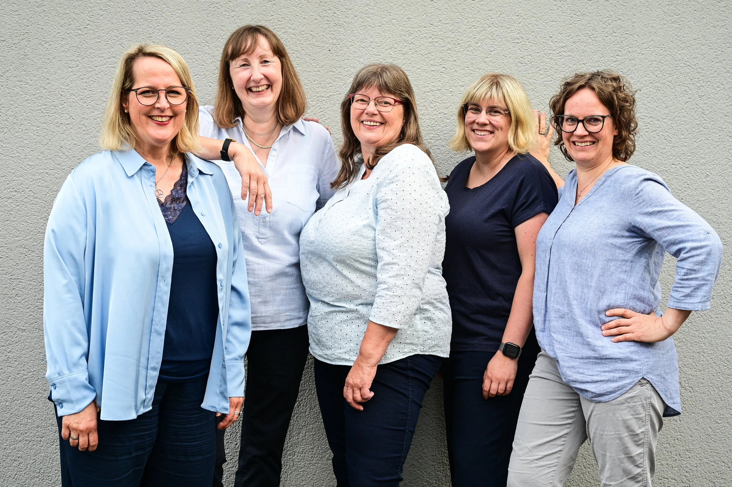 Sechs lachende Frauen stehen vor einer grauen Wand, tragen lässige Kleidung. Sie wirken fröhlich und entspannt.