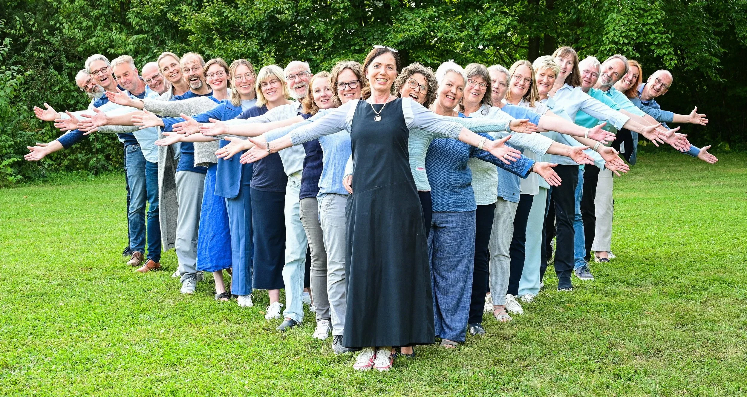 Gruppe von Menschen steht im Freien auf einer Wiese, alle strecken die Arme seitlich aus, lachend und freundlich lächelnd.