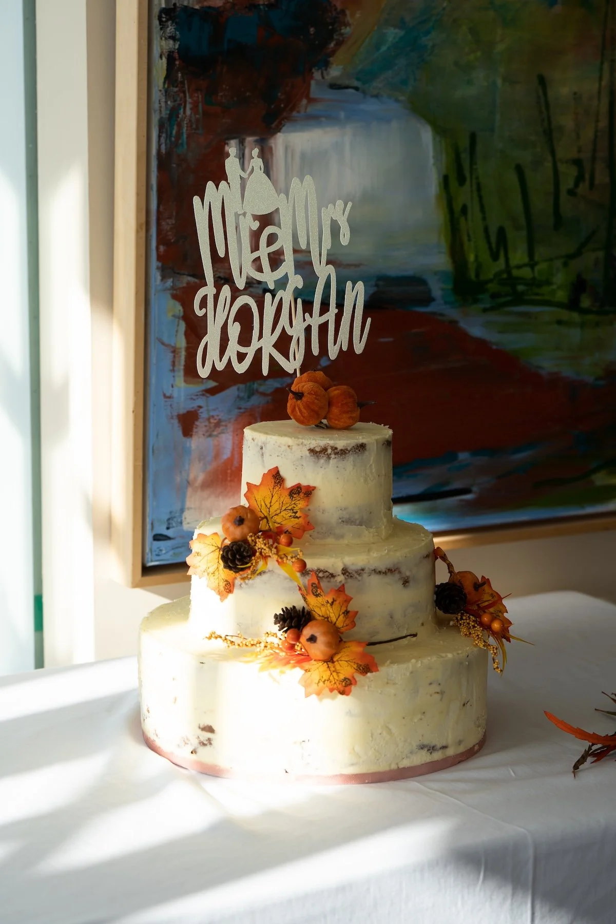 Three-tiered wedding cake decorated with fall leaves, berries, pinecones, and dried fruits, with a wedding topper reading 'Mr. & Mrs. Jackson' on a table with a white cloth, against a colorful abstract painting.