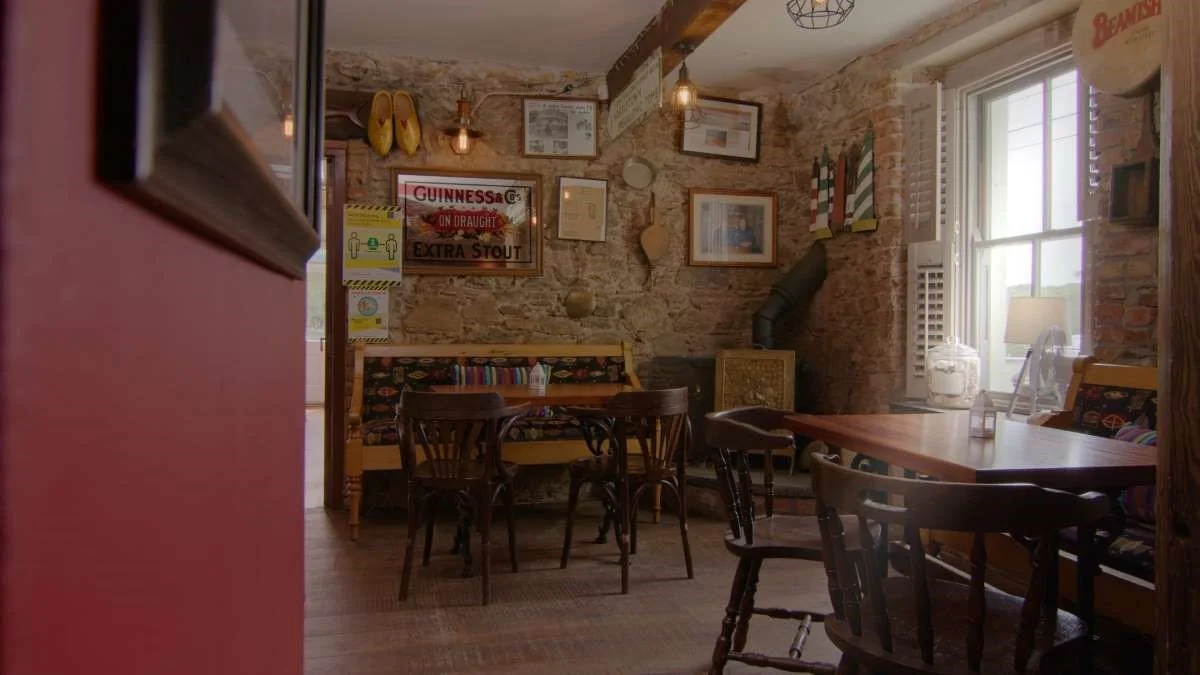 Interior of a cozy restaurant or pub with wooden furniture, brick walls decorated with framed pictures, lamps, and vintage signs, including a Guinness poster, with natural light from a window.