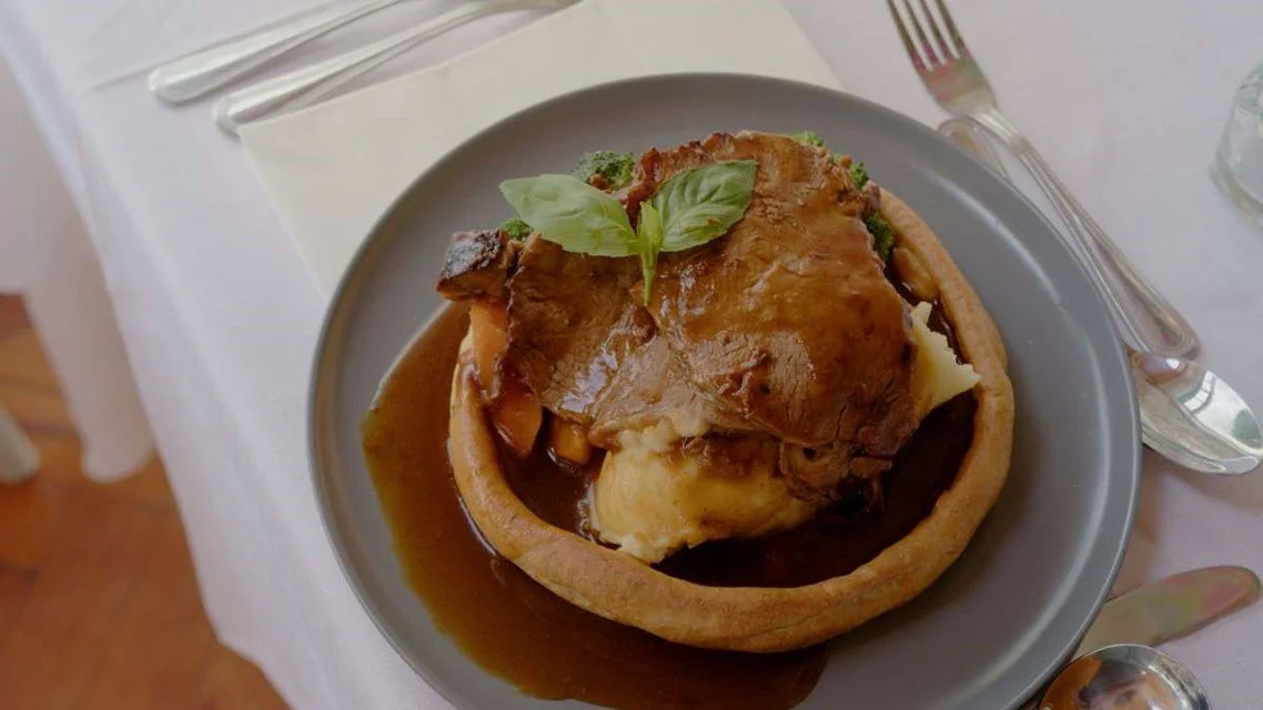 A savory meat pie topped with gravy and garnished with fresh basil leaves on a gray plate, set on a table with cutlery and a white tablecloth.
