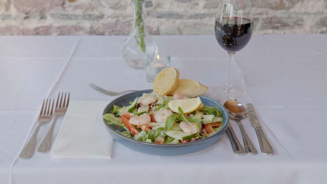 A seafood salad with shrimp, lettuce, tomato, and lemon slices in a gray bowl, accompanied by a glass of red wine on a white tablecloth, with forks and knives set on napkins.