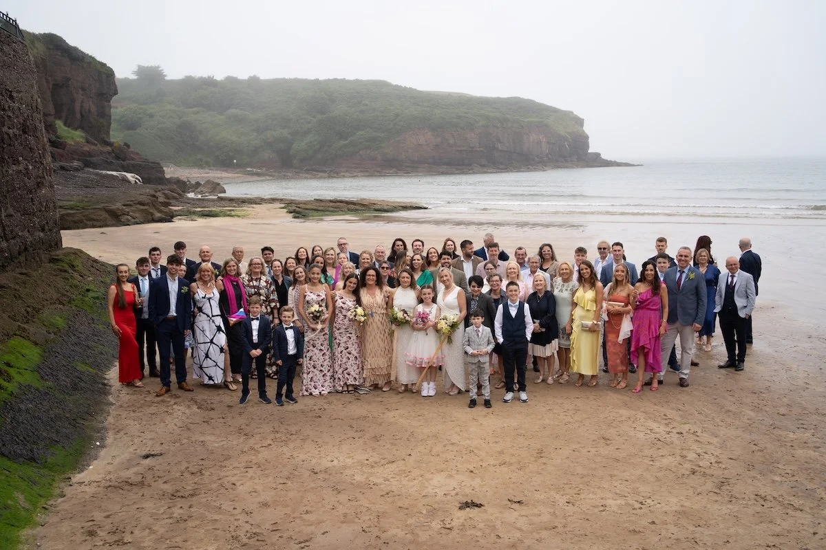 Group of people at a beach during a wedding ceremony, with cliffs and ocean in the background.