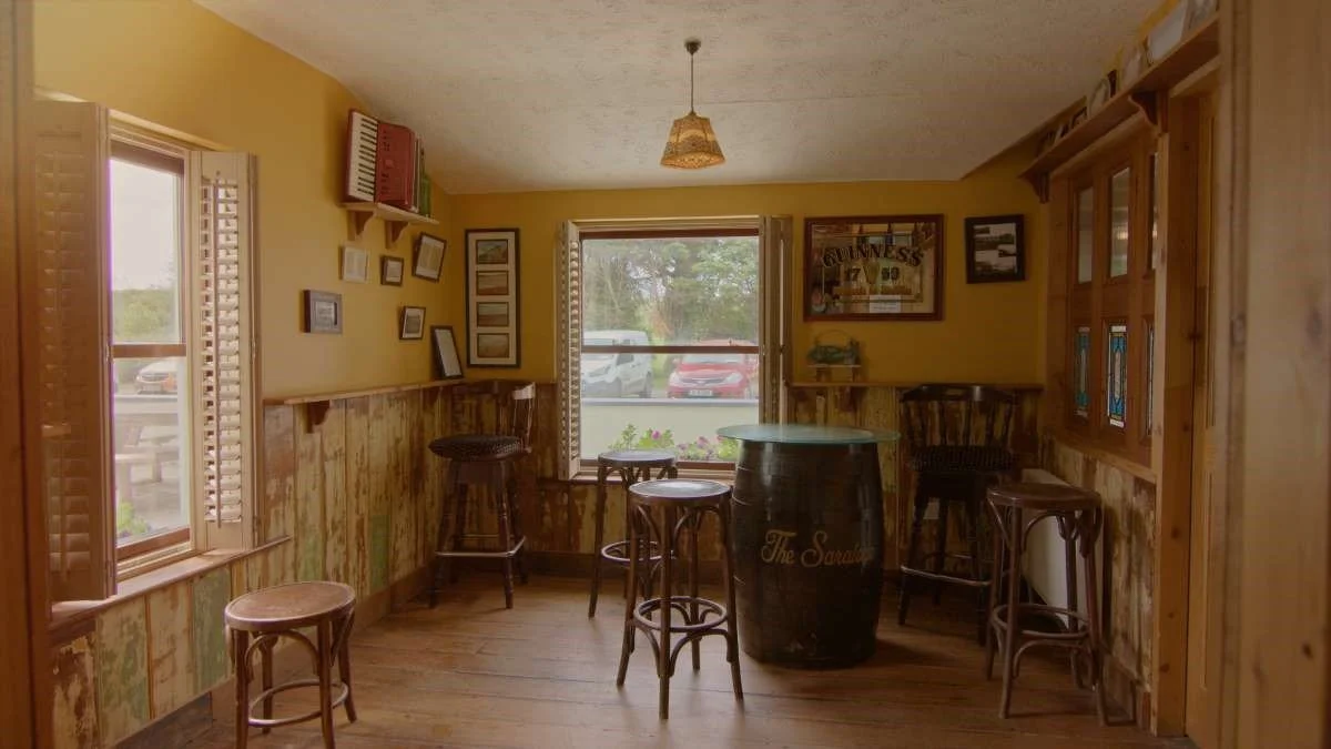Cozy bar area with yellow walls, wooden barstools, a barrel table, framed pictures, and a window view of parked cars outside.