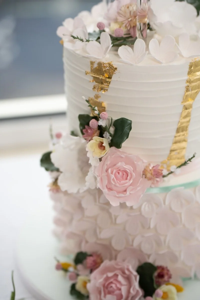 A close-up of a wedding cake decorated with pink and white flowers, green leaves, and gold accents.