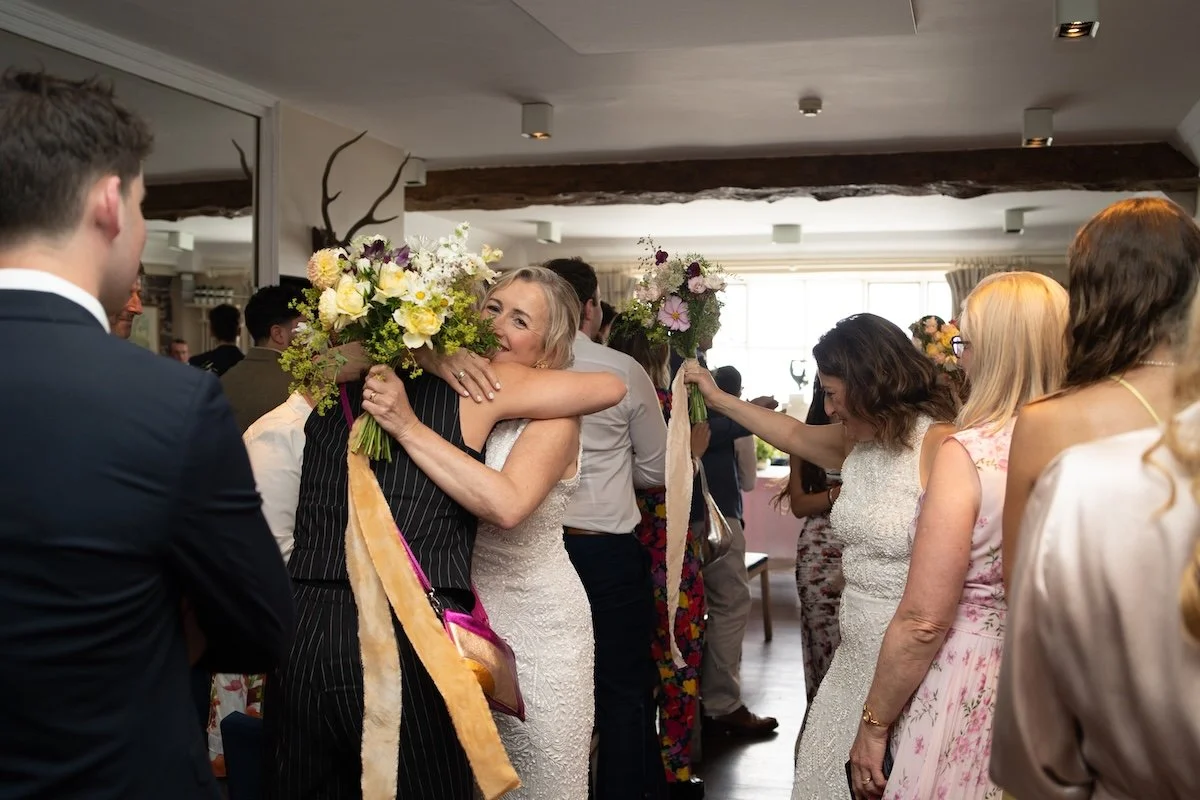 People celebrating at a wedding reception, hugging, and holding floral bouquets in a decorated indoor venue.