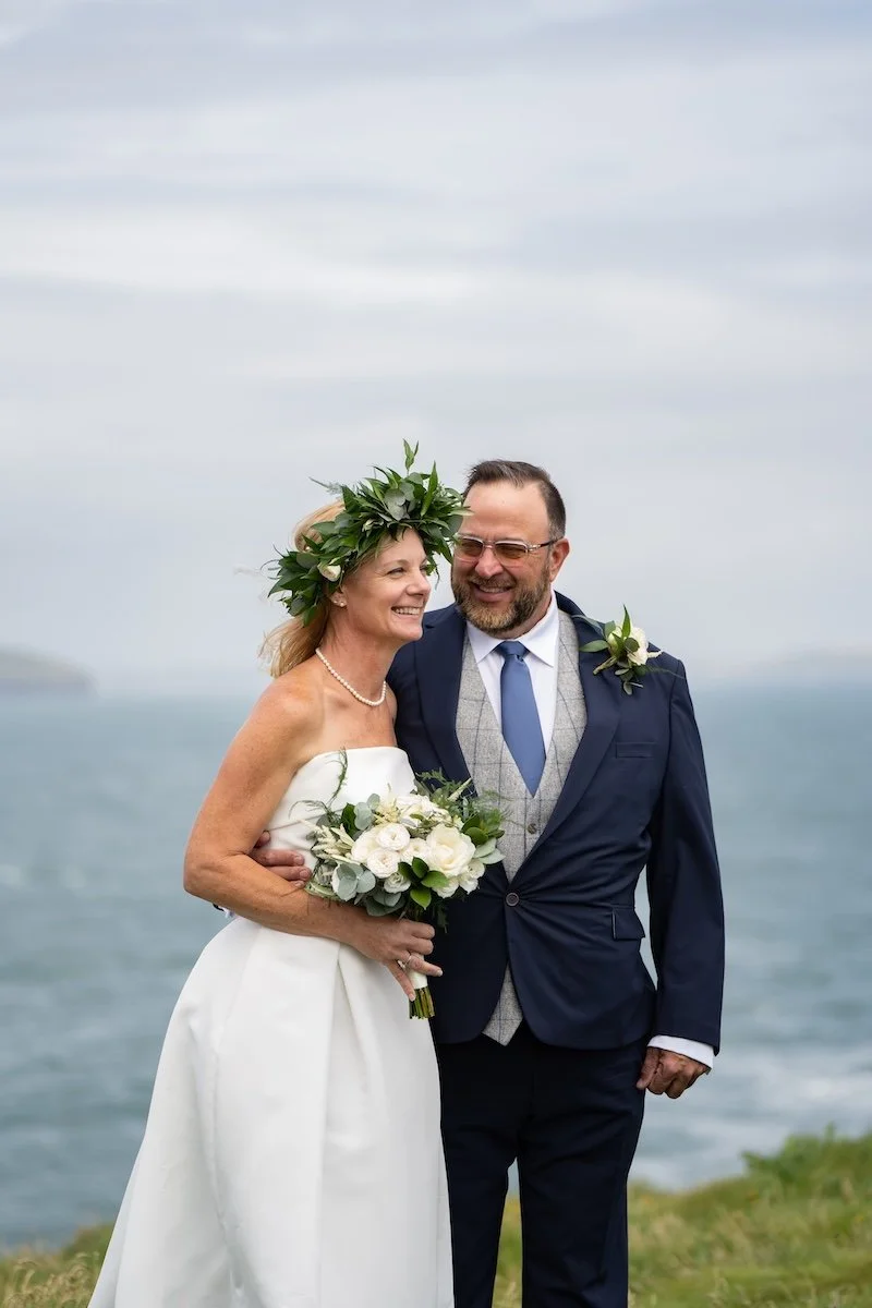 A couple dressed in wedding attire, standing outdoors near the ocean, smiling and holding a bouquet of flowers, with a cloudy sky in the background.