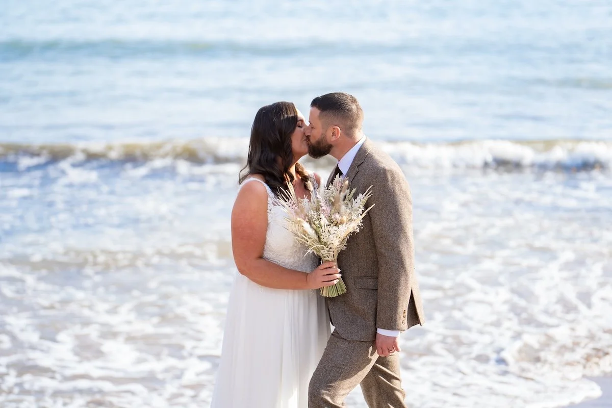 A newlywed couple sharing a kiss on the beach with ocean waves in the background. The bride is holding a bouquet of flowers, wearing a white dress. The groom is dressed in a light gray suit.