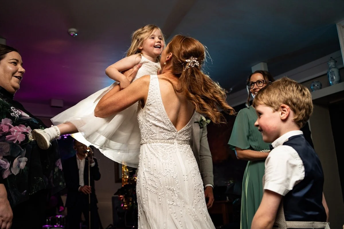 A woman in a wedding dress holding a young girl dressed in a white dress, lifting her up and smiling. Several adults, including a woman in glasses, and a young boy in a vest and bow tie, are observing and smiling at the interaction.