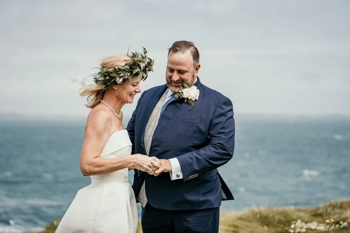 A couple dressed in wedding attire holding hands and smiling near the ocean, with a cloudy sky above.