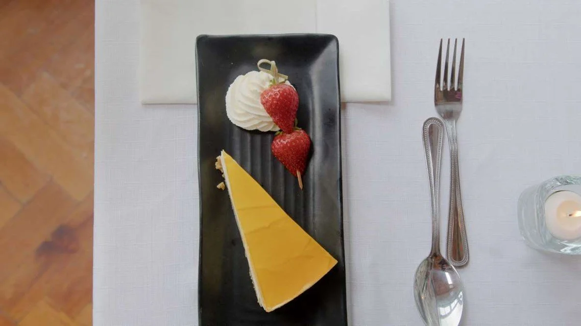 Slice of lemon cheesecake, whipped cream topped with strawberries, and a decorative chocolate piece on a black rectangular plate, with a fork, a white napkin, and a lit candle on a white tablecloth
