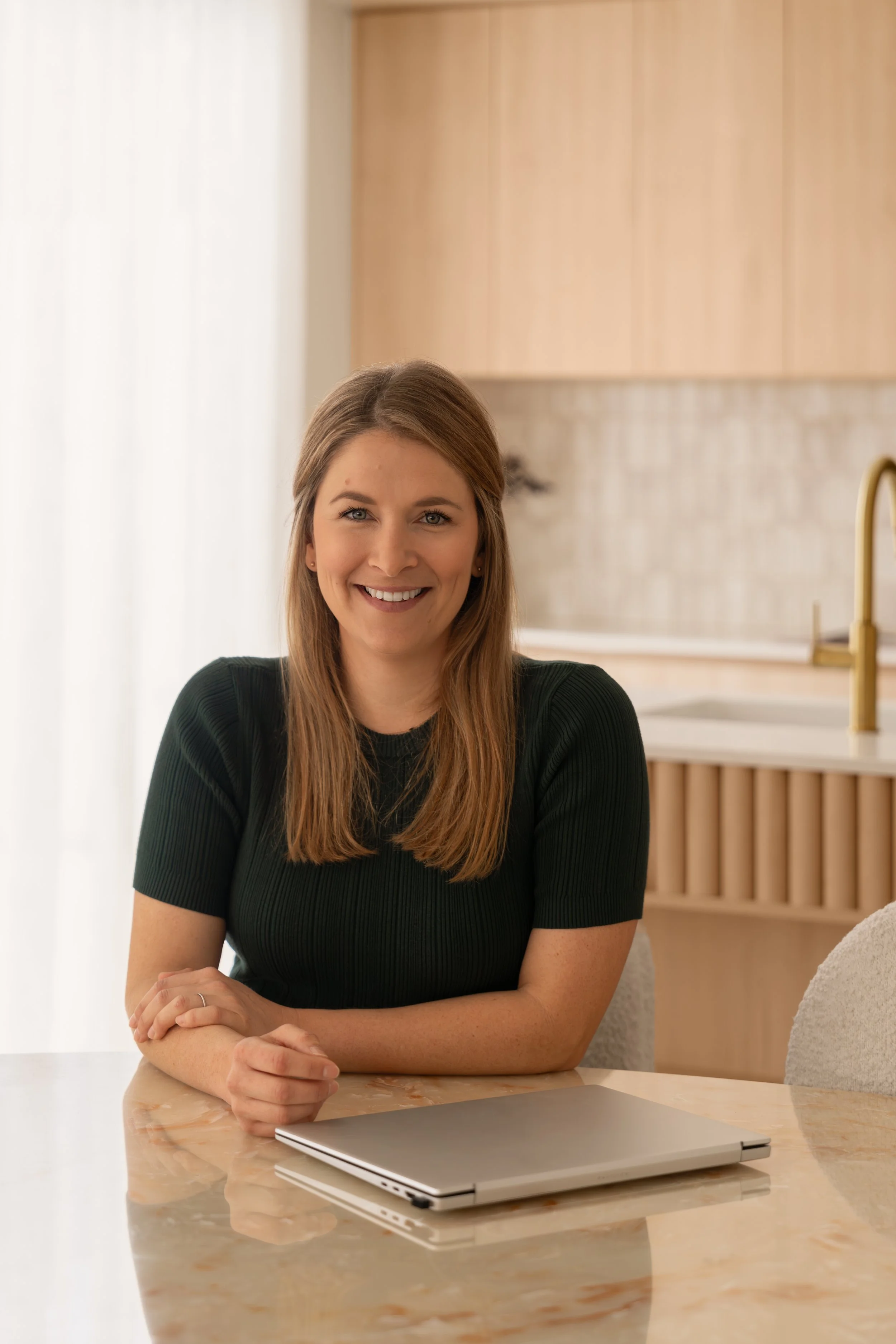 A woman seated at a marble table in a brightly lit modern kitchen, with her arms resting on the table and a laptop in front of her, smiling at the camera.