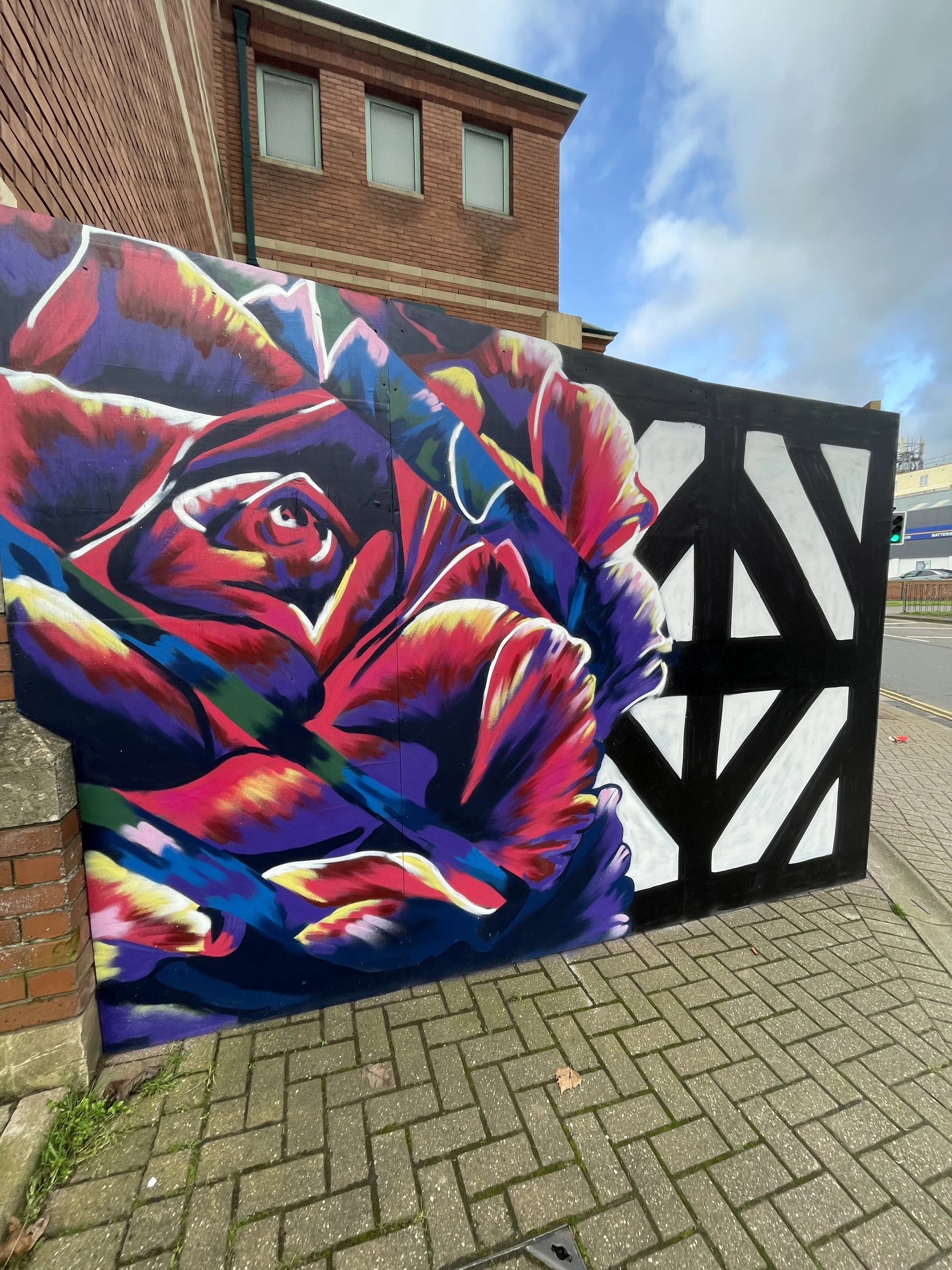 A colorful mural painted on a wall shows a stylized face of an elderly man with glasses and a beard, painted in bright reds, purples, blues, and yellows. The mural is situated outdoors on a city sidewalk with brick and wood buildings nearby.