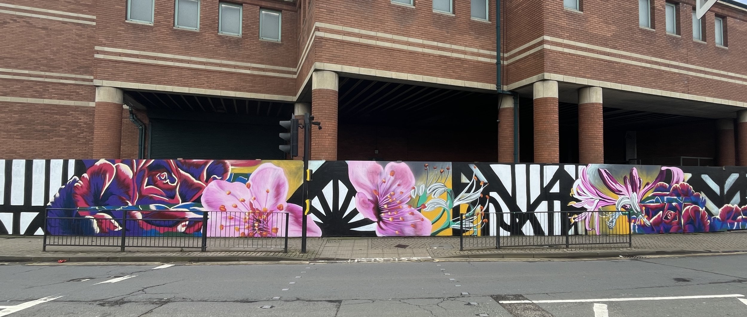 A colorful mural painting of pink flowers, including lilies and roses, on a wall in front of a brick building with arched and rectangular windows.