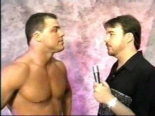 Two men standing face to face, one shirtless and one holding a microphone, against a pinkish textured background.