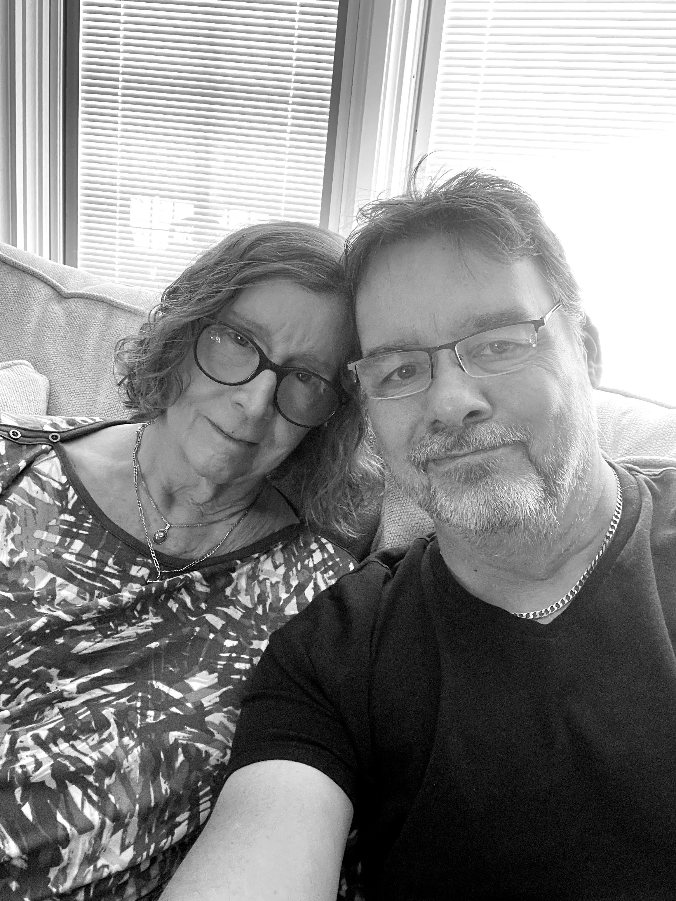 A black and white selfie of a man and woman sitting on a couch, both wearing glasses. The woman has shoulder-length hair and is wearing a patterned top, while the man has short hair and a beard, wearing a black t-shirt. They are close together, smiling softly, with window blinds in the background.