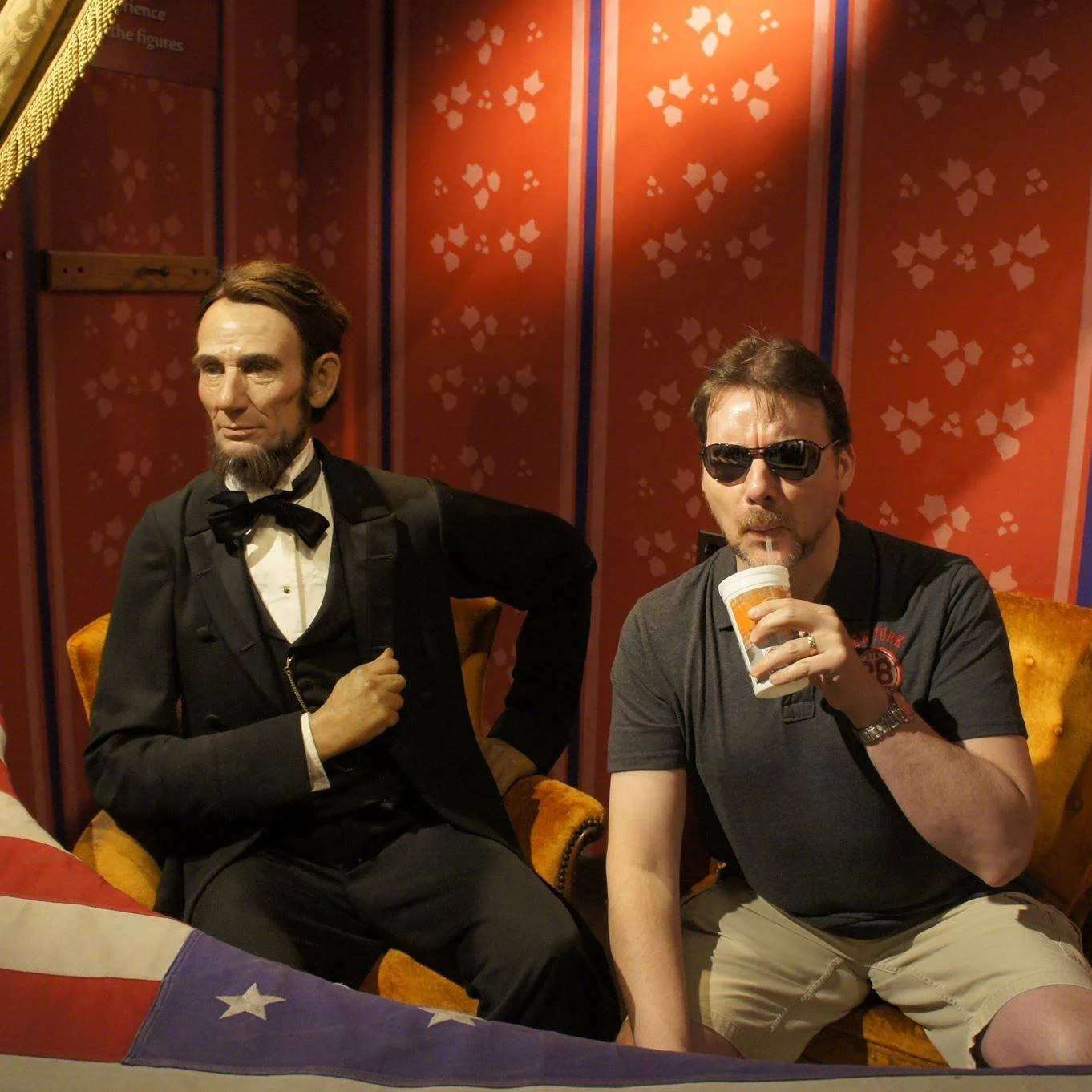 A man wearing sunglasses and a black t-shirt sitting on an orange vintage chair, drinking from a cup with a straw. Next to him is a wax figure of Abraham Lincoln dressed in a formal black suit with a bow tie, seated on a similar vintage chair. The ba