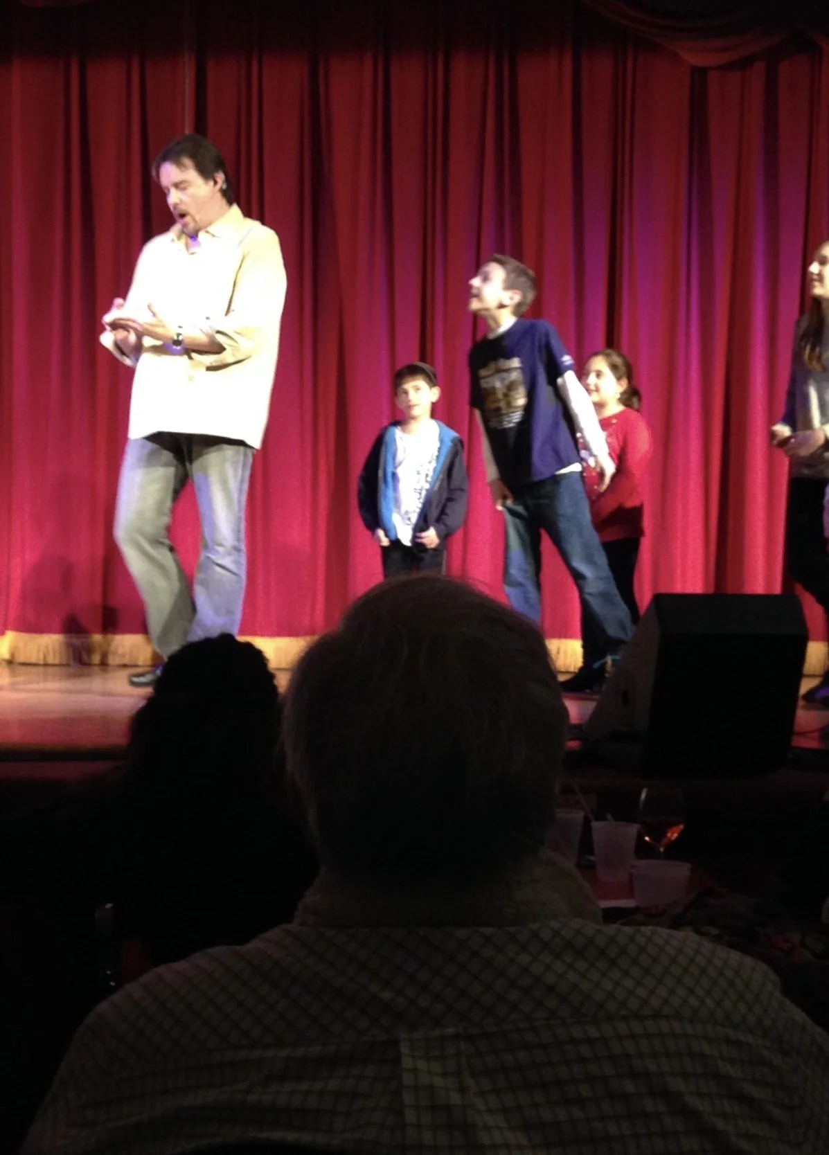 A man with dark hair, wearing a light-colored shirt and gray pants, standing on a stage with a red curtain, appears to be speaking or presenting. Four children stand behind him, looking at him or the audience. Audience members are visible in the foreground.