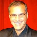 A smiling man with glasses and short gray hair, wearing a dark suit jacket, against a red background.