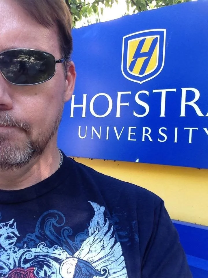 A man wearing sunglasses and a graphic T-shirt taking a selfie next to a blue sign for Hofstra University.
