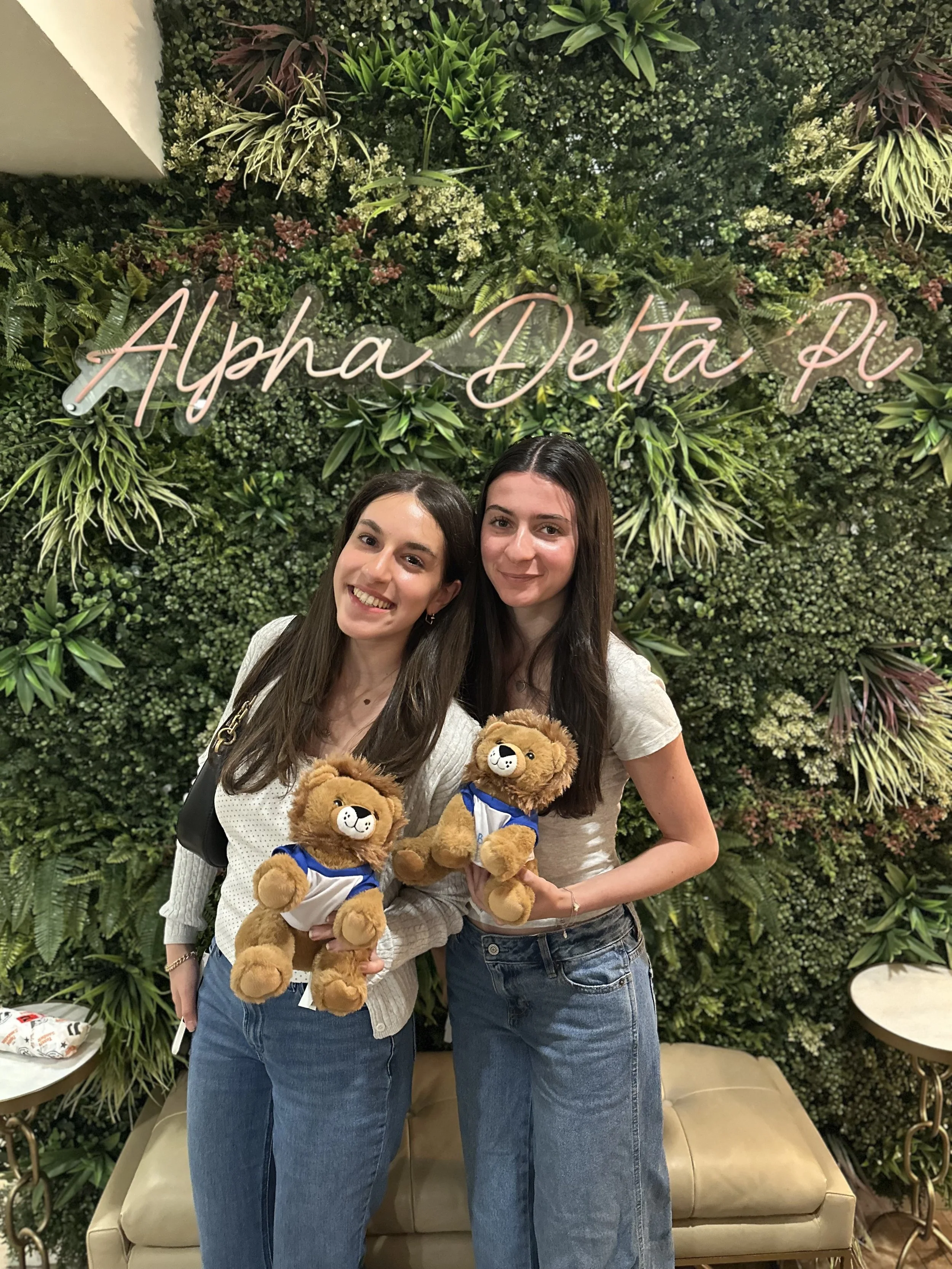 Two women holding plush lion toys, standing in front of a green wall with a neon sign that reads 'Alpha Delta Pi.'